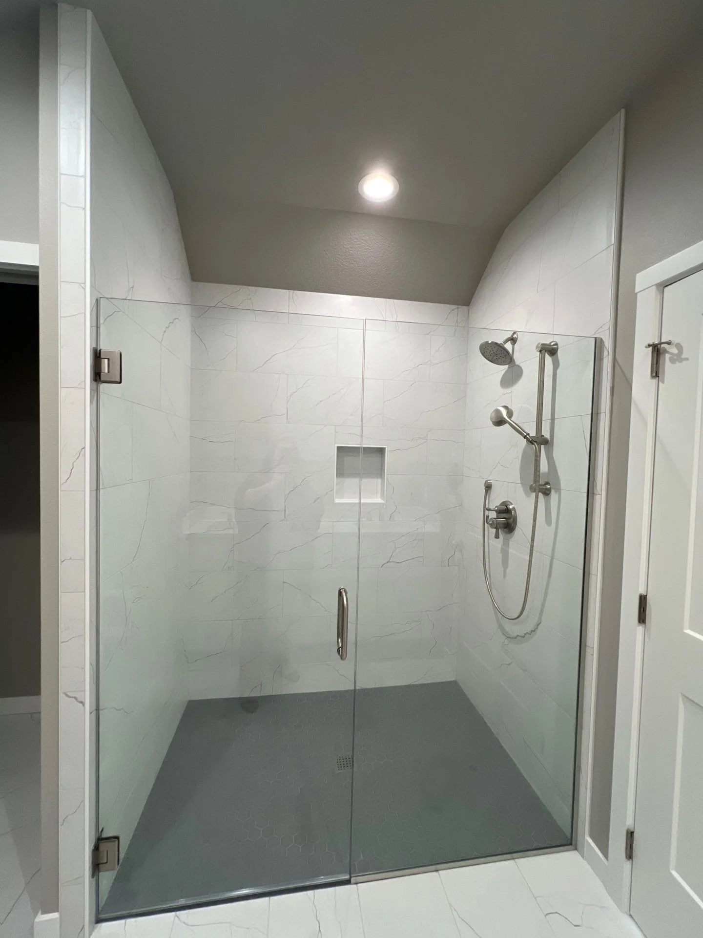 Modern walk-in shower with glass door, white marble tile walls, gray hexagonal tile floor, built-in niche, and chrome shower fixtures.