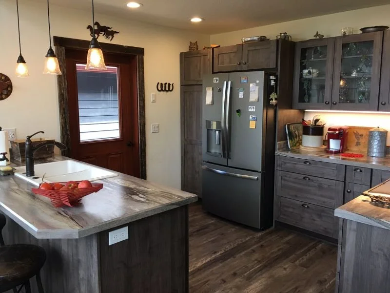A kitchen with dark wooden cabinets, a stainless steel refrigerator, and a marble countertop island with a bowl of apples. Hanging pendant lights above the island, a window with a wooden door nearby, and various kitchen items and decorations.