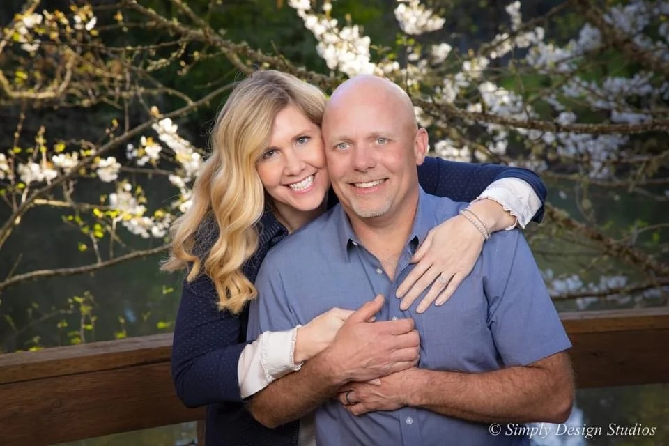 A smiling woman hugging a bald man from behind outdoors, with blooming tree branches in the background.