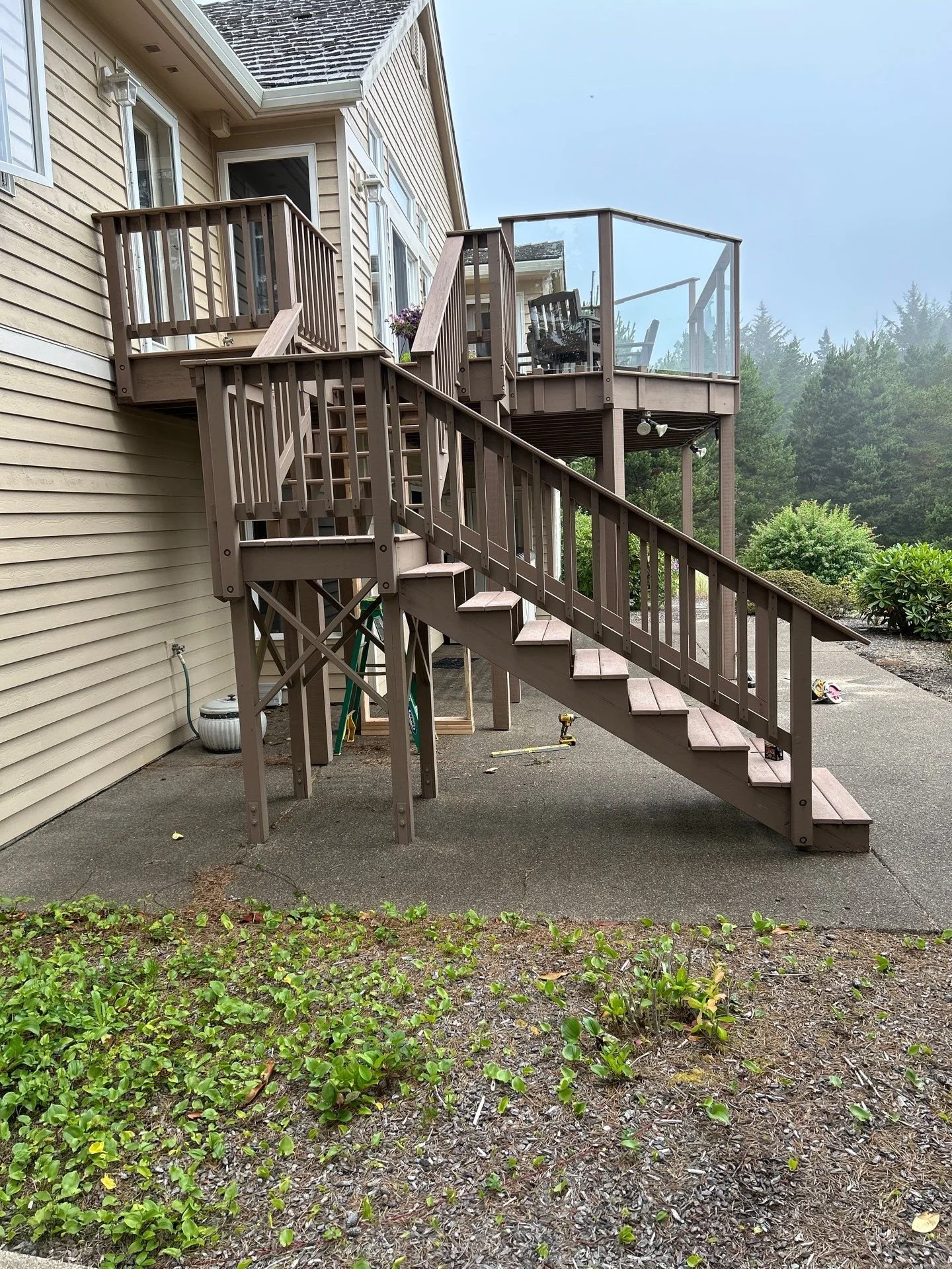 A multi-level outdoor wooden deck with stairs attached to beige house siding, surrounded by a garden and forested area in the background.