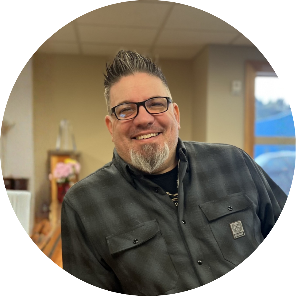 A smiling man with glasses and a beard, wearing a plaid shirt, standing indoors near a window.