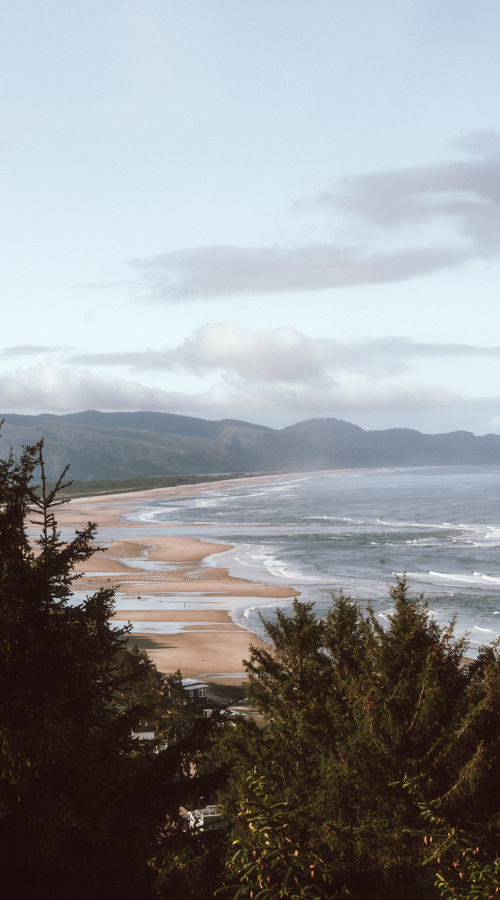 A scenic view of the Central Oregon Coast with sandy beaches, ocean waves, and a backdrop of mountains under a partly cloudy sky, with trees in the foreground.