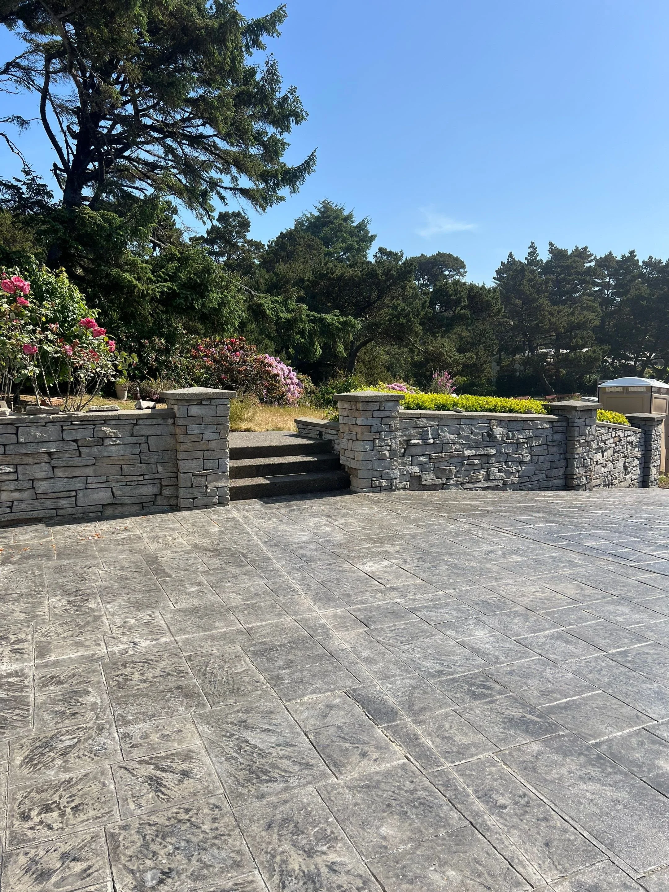 A stone patio with stairs leading up to a raised garden area, surrounded by a stone wall, with trees and flowering bushes in the background under a clear blue sky.