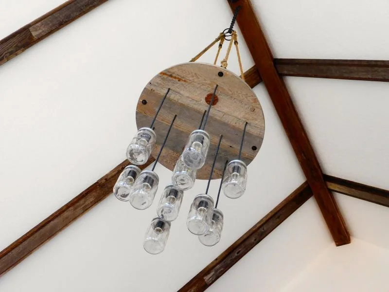 A rustic chandelier made from a large wooden disc and upside-down mason jars, hanging from the ceiling with a rope and pulley system.