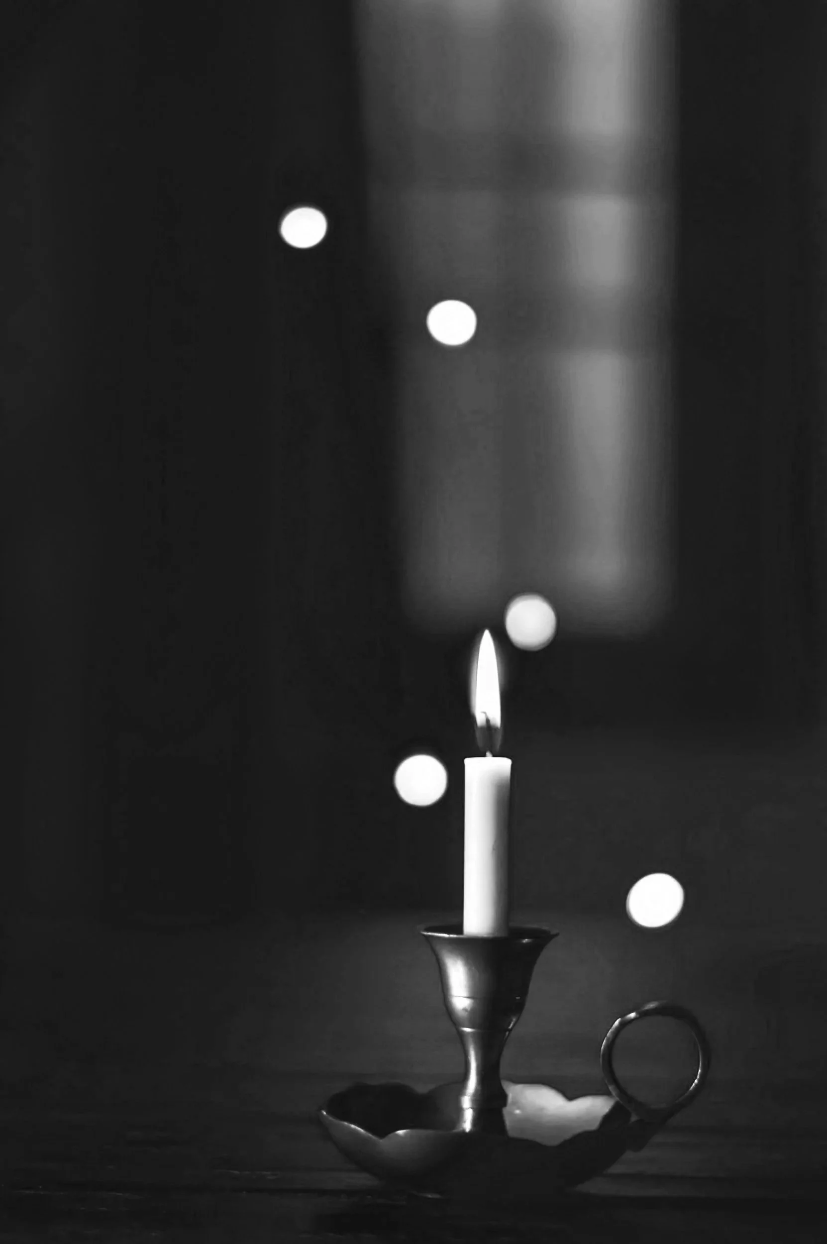 A lit candle in a metal holder, with blurred falling snow or dots in the background, in black and white.