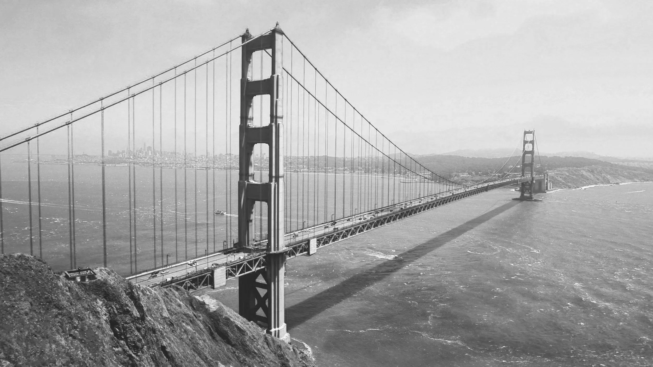 Black and white photo of the Golden Gate Bridge spanning over water, with a city skyline in the background.