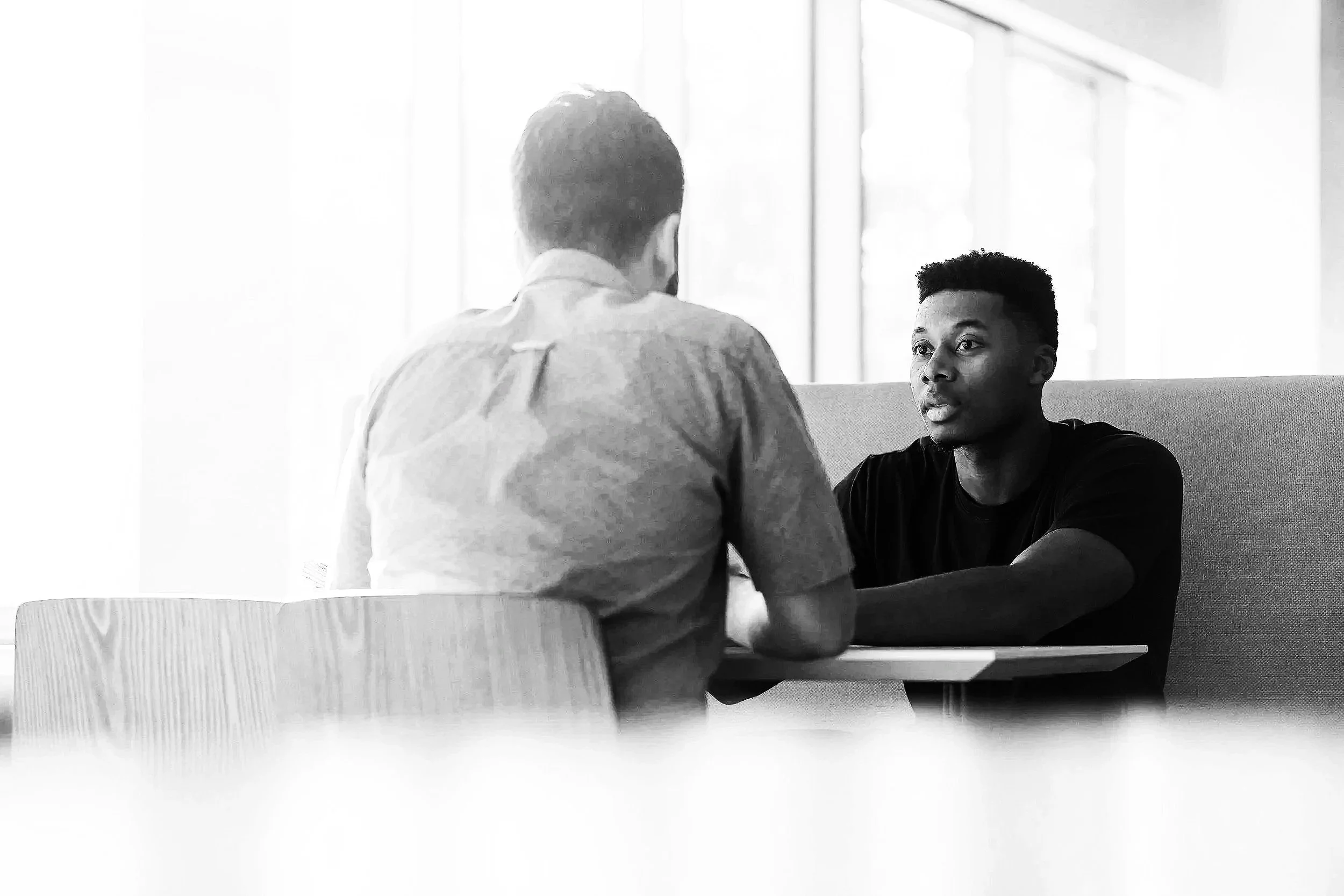 Two men engaged in a serious conversation at a table in a well-lit room with large windows. One man, facing away, is wearing a light-colored shirt; the other, facing forward, is wearing a dark shirt.