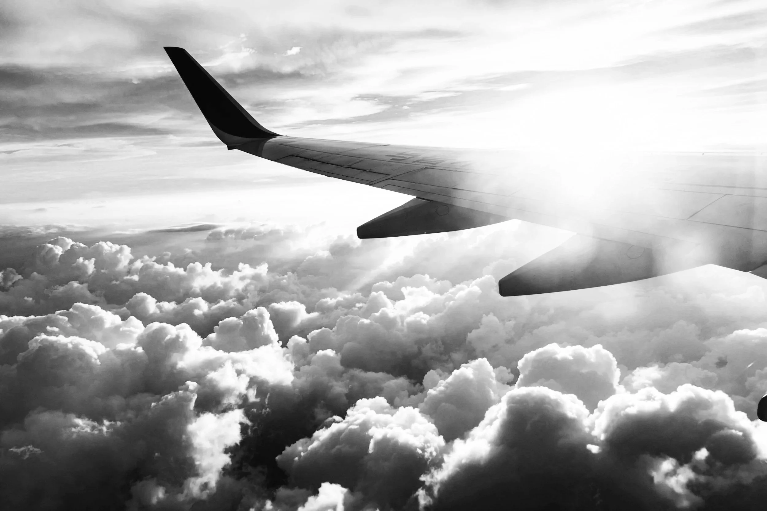 Black and white photo of an airplane wing flying above clouds during sunset or sunrise.