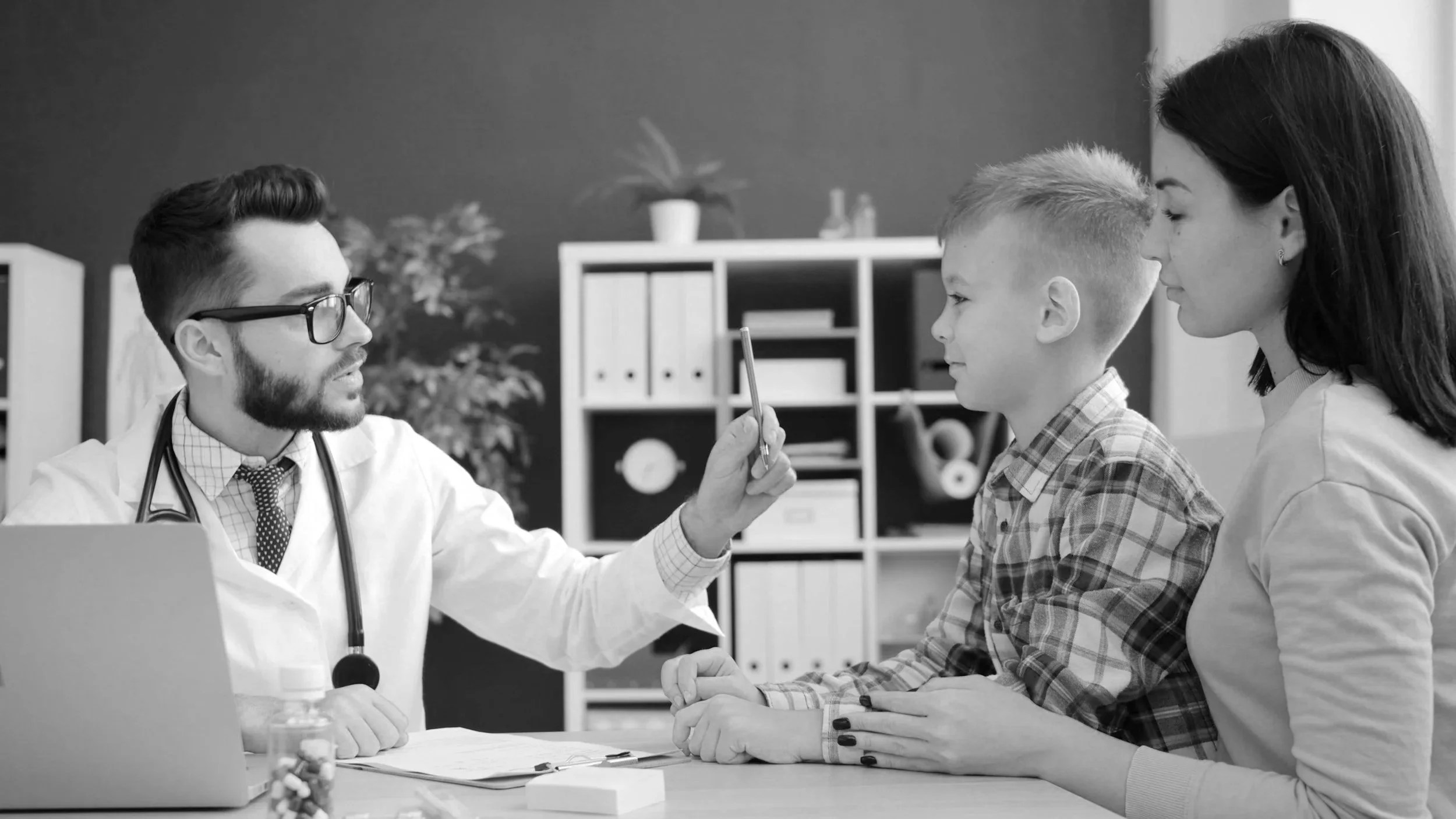 A doctor in a white coat and glasses is speaking to a young girl and her mother during a medical consultation in an office. The doctor is holding a pen and gesturing as he talks, with a laptop and bottles on the table.