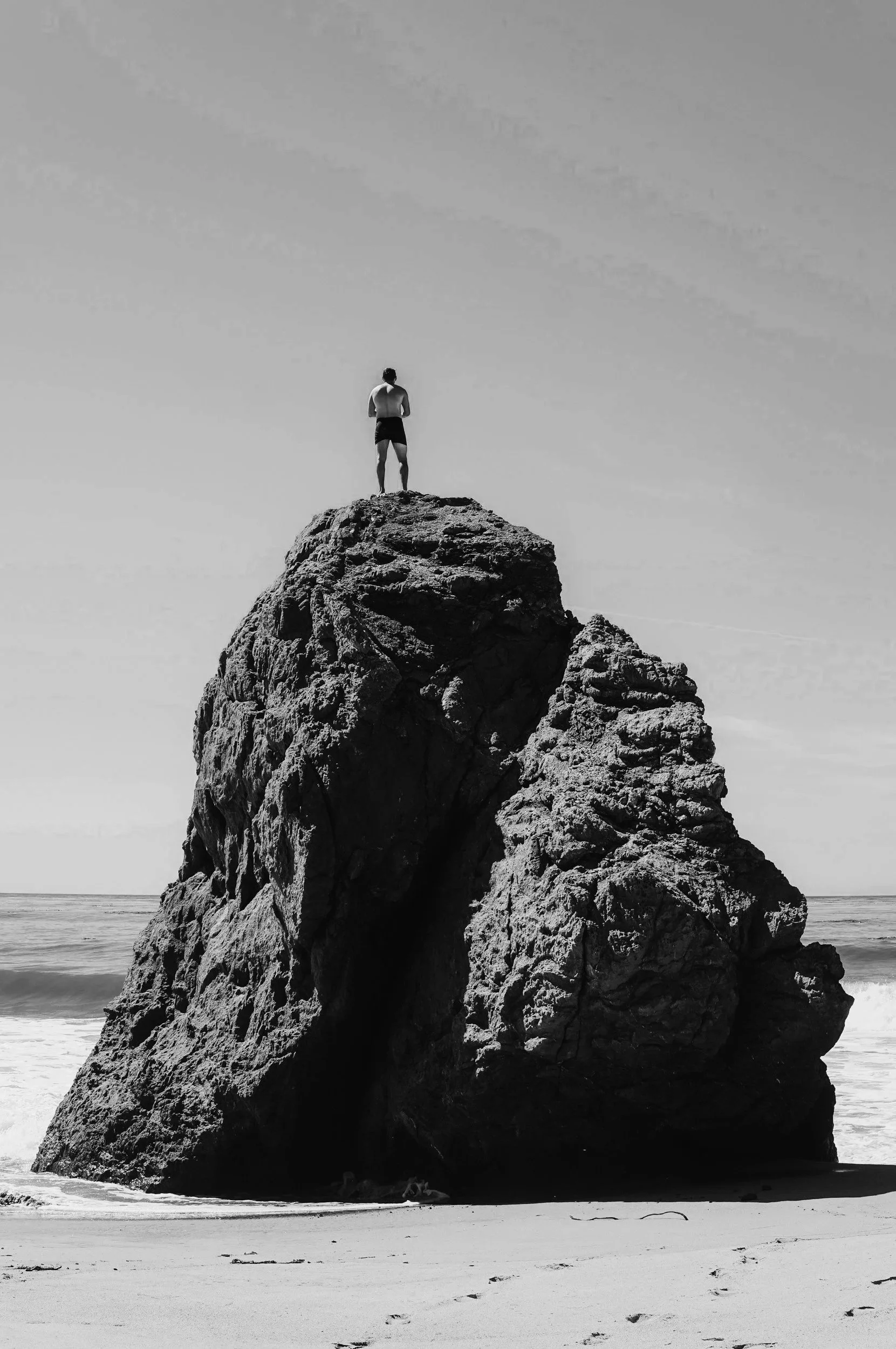 A person standing on top of a large rock formation at the beach, facing the ocean, in black and white.