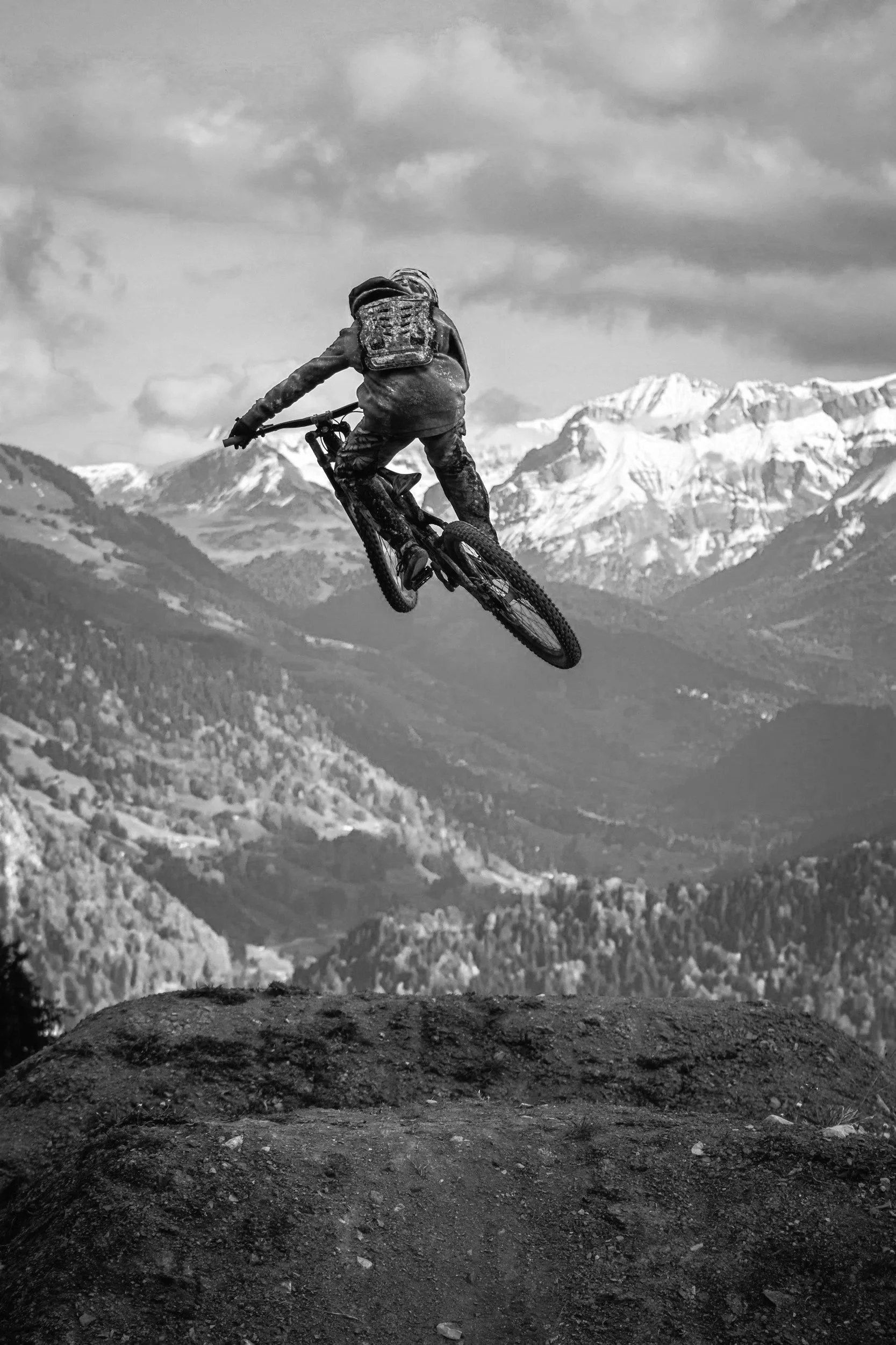 A person riding a mountain bike and jumping off a dirt ramp in a mountainous landscape with snow-capped peaks in the background.