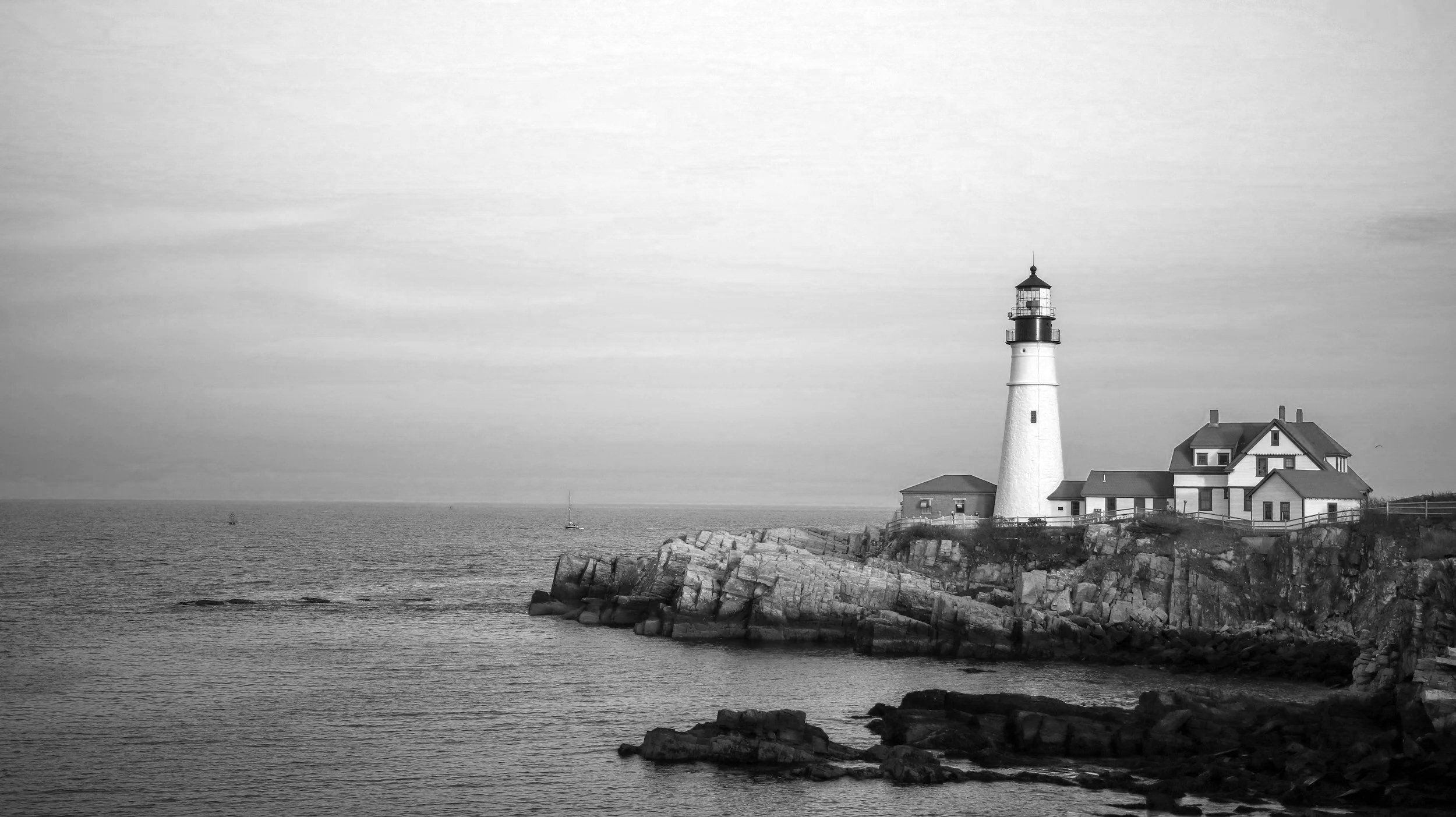 A black and white photograph of a lighthouse on a rocky coastline with a house nearby and sailboats in the distance on the ocean.
