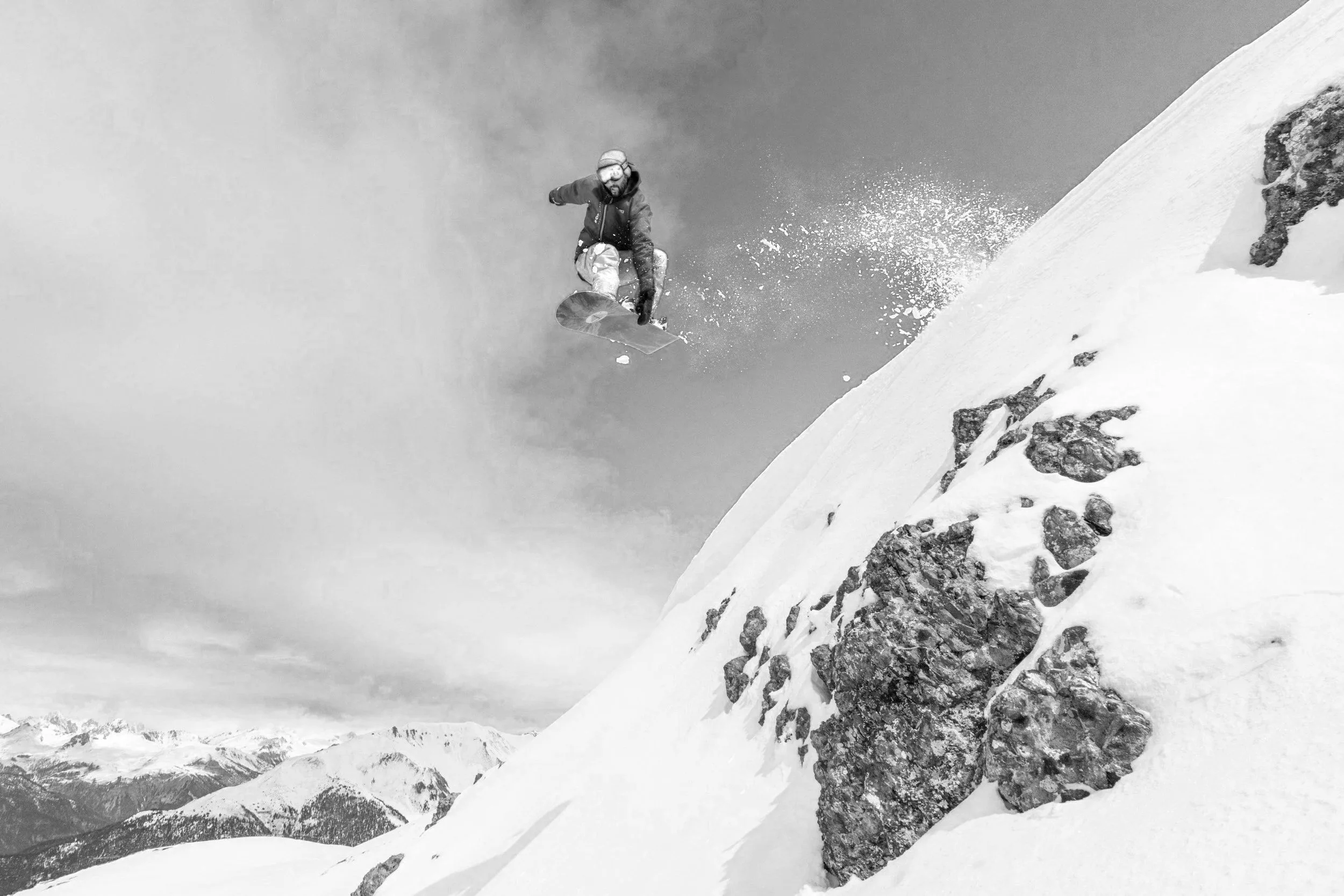A snowboarder in a jacket and helmet jumping off a snowy mountain slope with rocky outcroppings, snow-capped mountains in the background, and a cloudy sky.