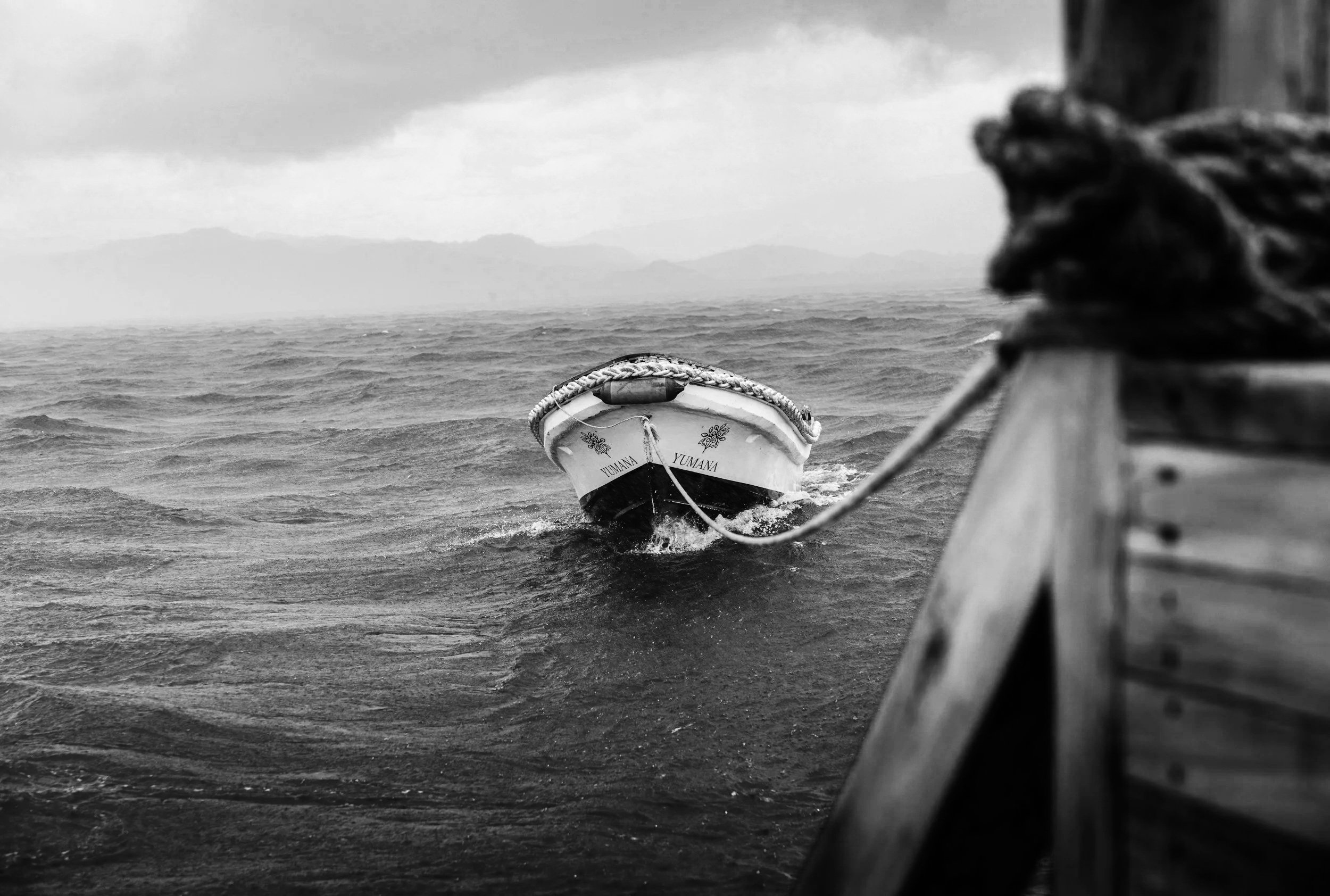 A boat named Yumana pulled away from a dock into choppy waters with mountains in the distance, depicted in black and white.