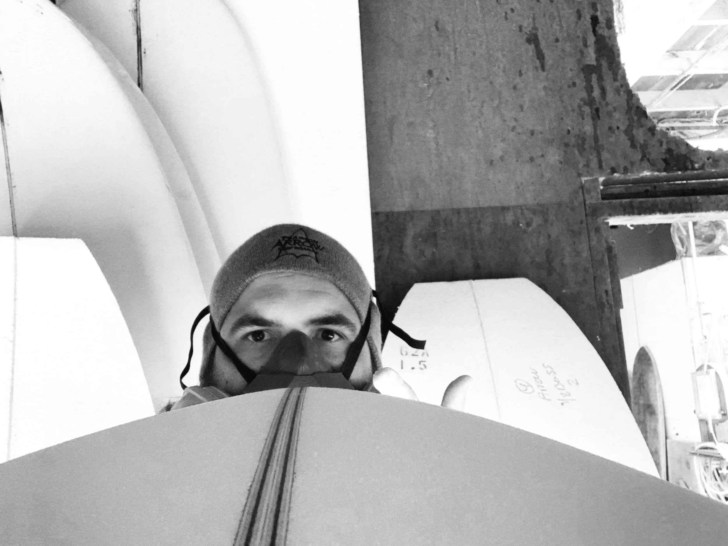 A person wearing a beanie and a mask, giving a thumbs-up, with a surfboard in front of them and a construction site or wooden surface background.