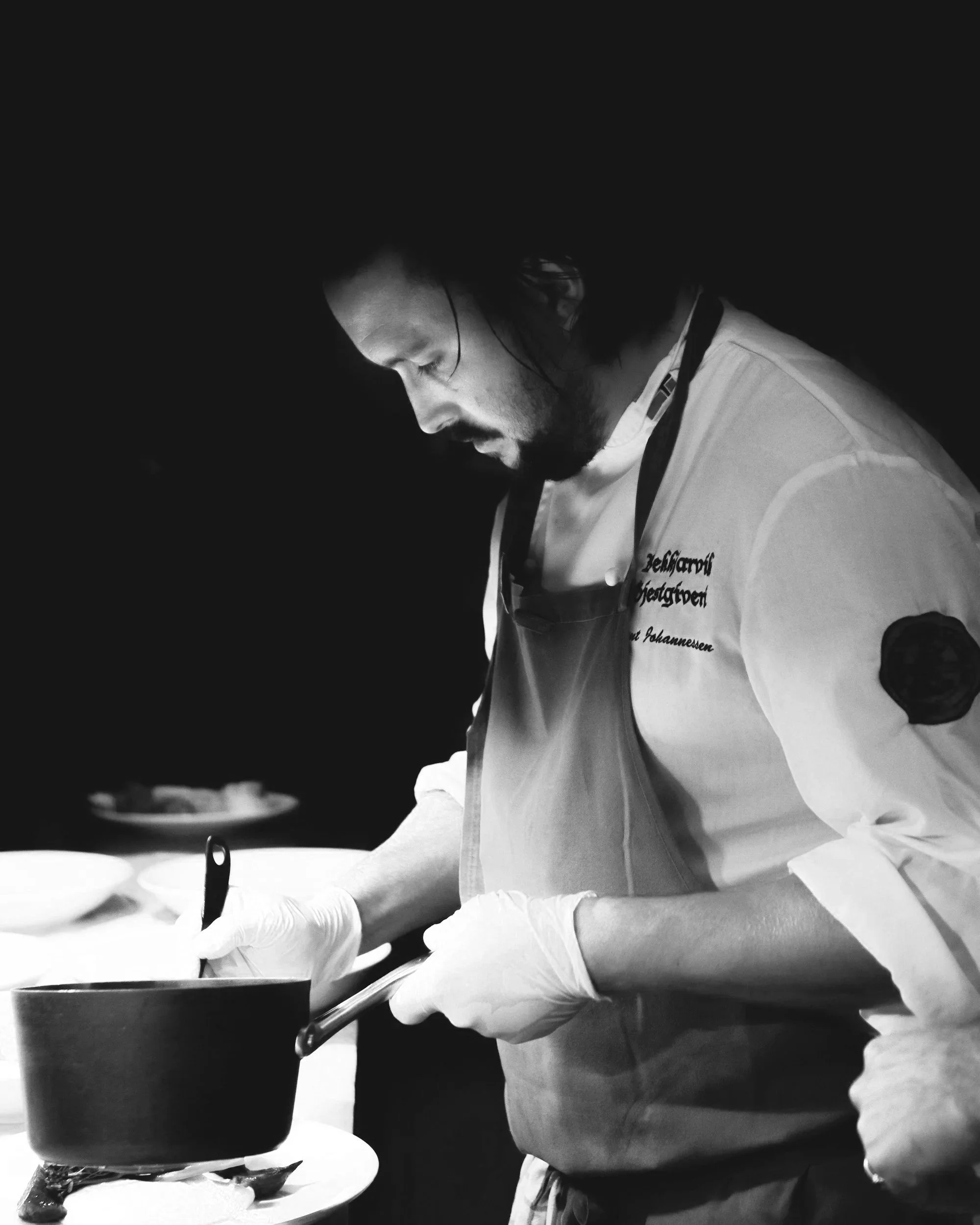 A chef wearing a white uniform and gloves, focused on cooking in a professional kitchen, with a dark background.