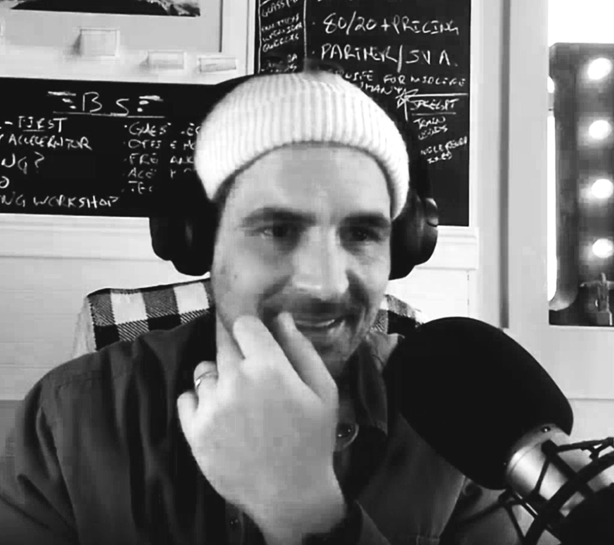 A man wearing a white knit cap and headphones, sitting in front of a microphone, with blackboards with writing on them in the background.