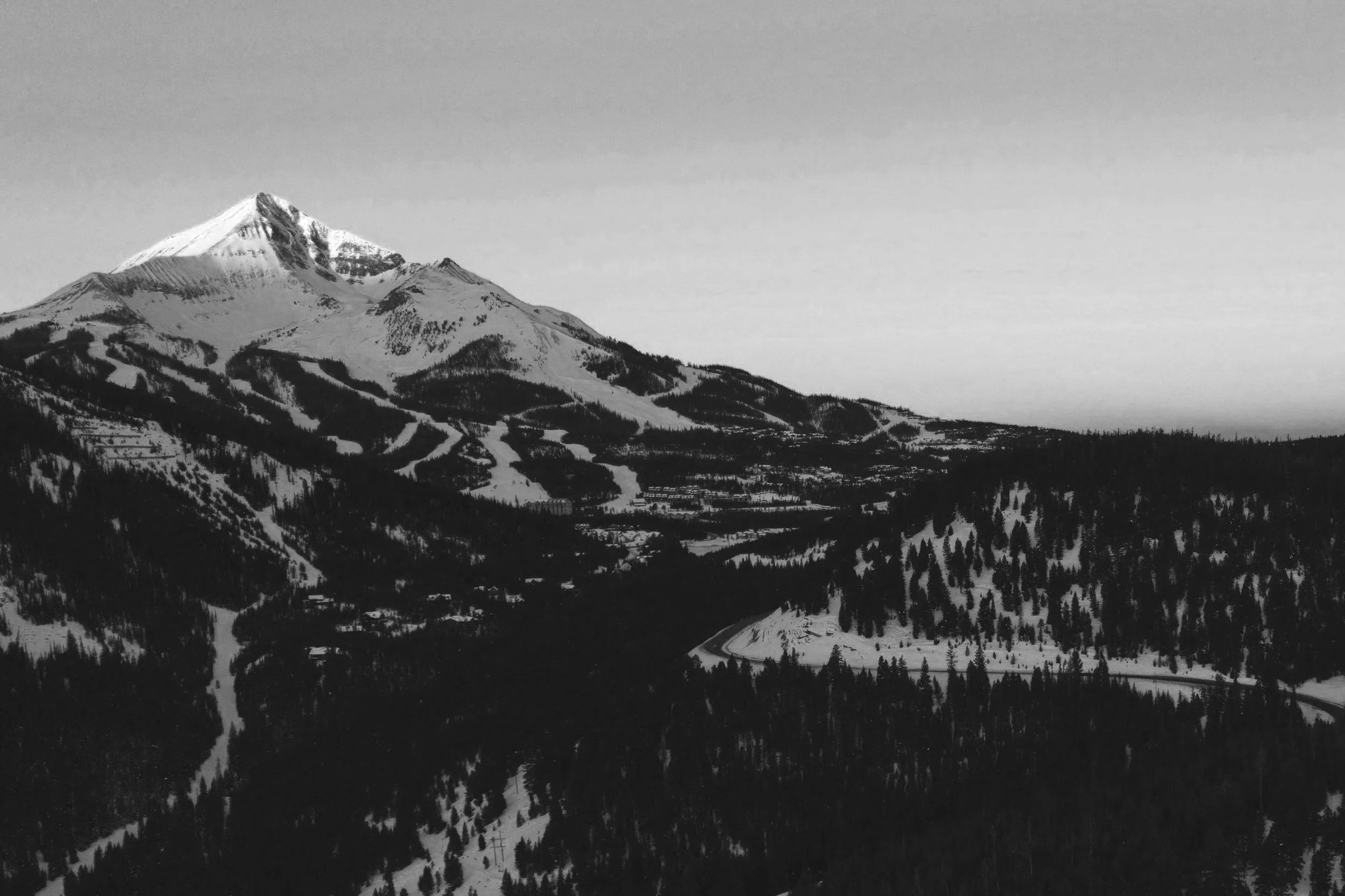 Black and white photo of snow-covered mountainous landscape with evergreen trees and winding roads.