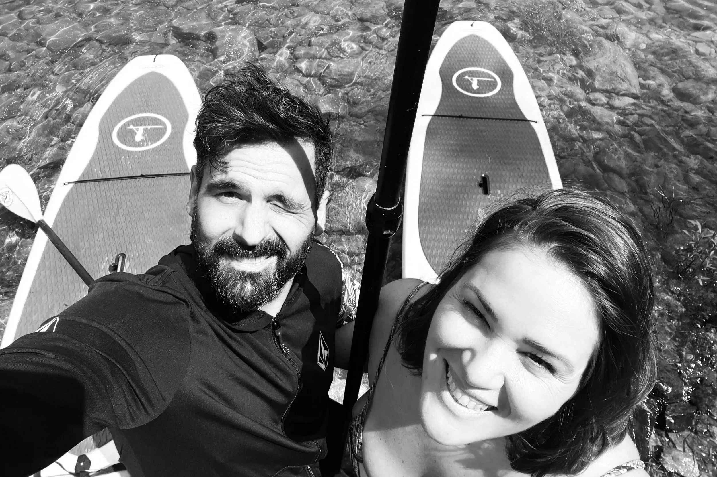 A man and a woman take a selfie on paddleboards in the water, with the man winking and smiling.