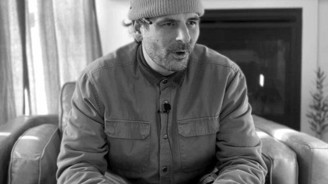 A man wearing a beanie and a button-up shirt, sitting on a sofa, talking or explaining with a serious expression. There is a television and a plant in the background.
