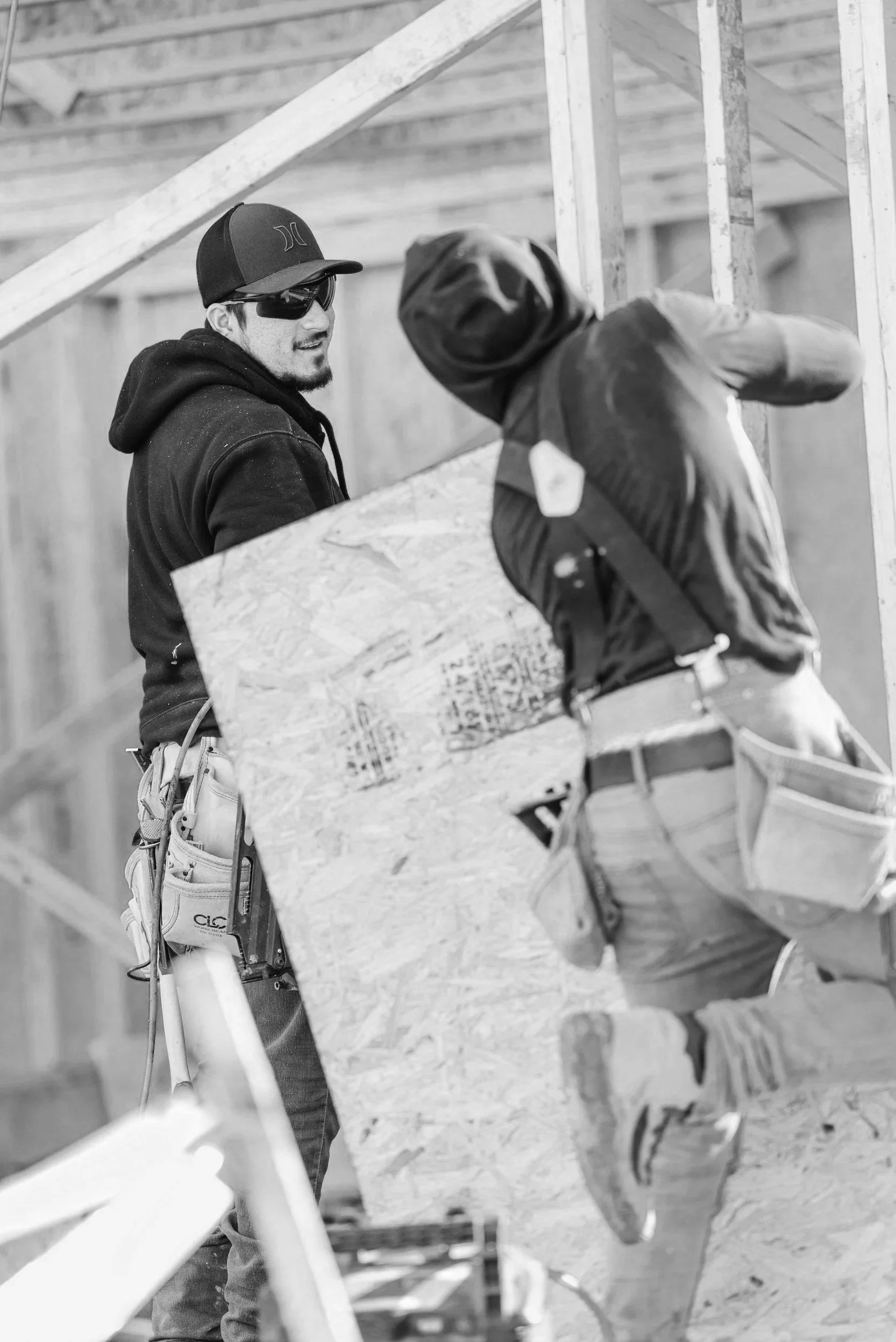 Two construction workers on a building site, one holding a wooden sheet and the other working on the frame of a structure.