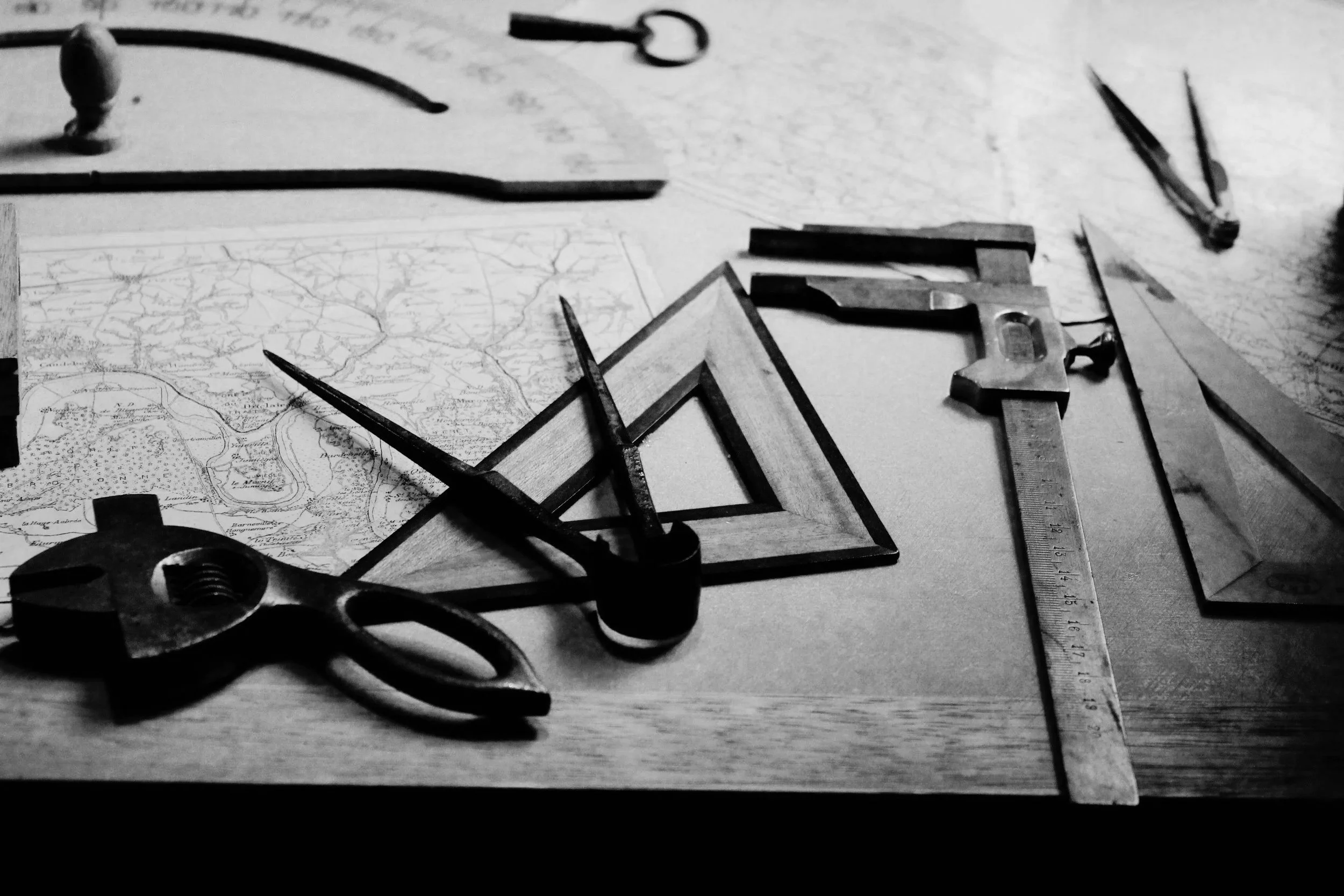 A black and white image showing drafting tools such as a compass, a straightedge ruler, a triangular ruler, and a frame surrounding a map on a wooden surface.