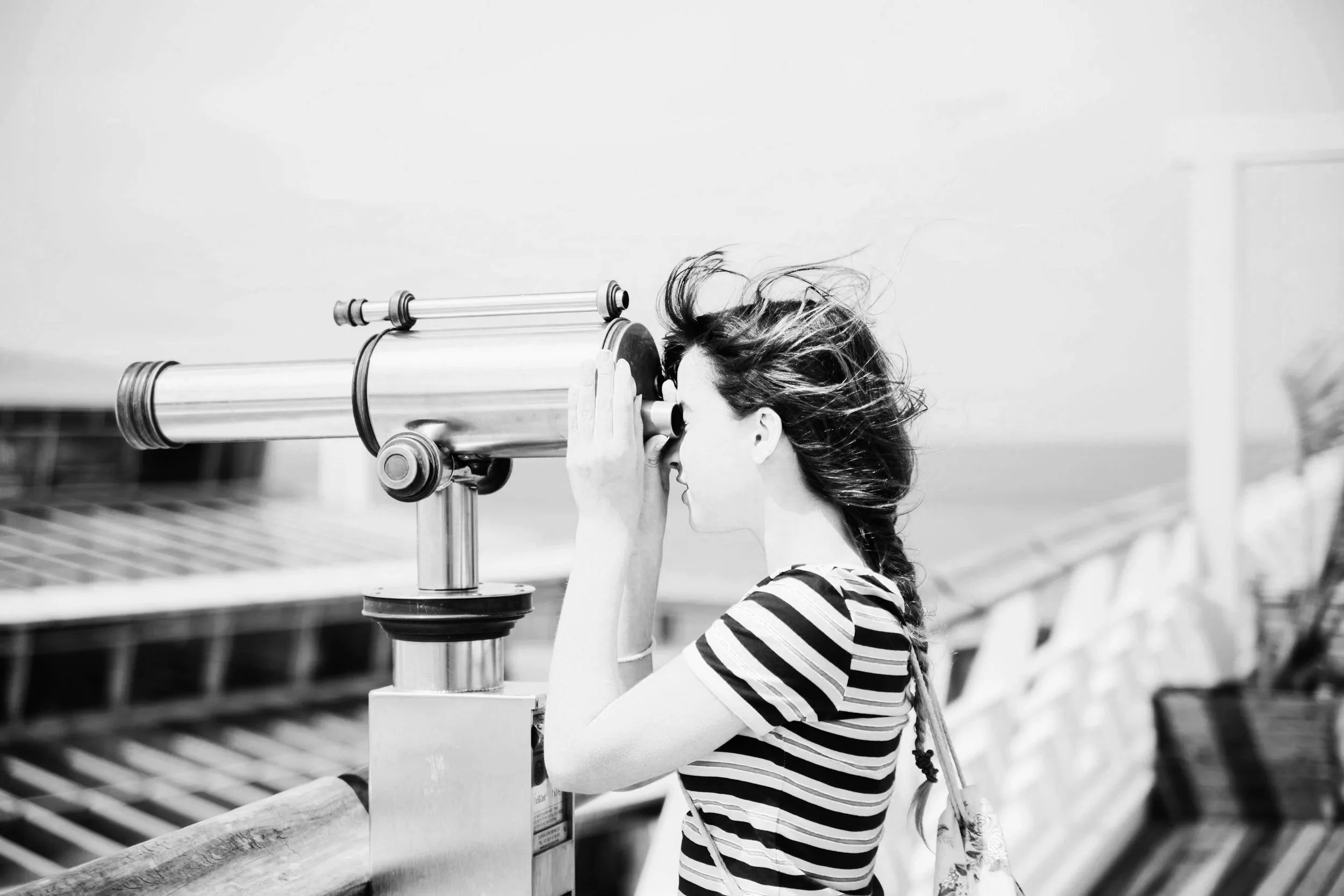 A woman with wind-blown hair looks through a coin-operated binocular viewer on a pier or outdoor observation deck.