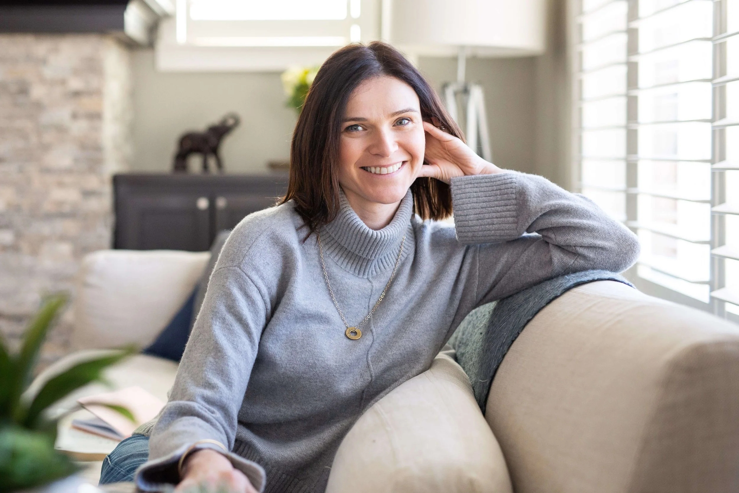A woman with shoulder-length dark hair, wearing a gray turtleneck sweater and a necklace, smiling and sitting on a beige sofa in a brightly lit living room with large windows and a brick wall in the background.