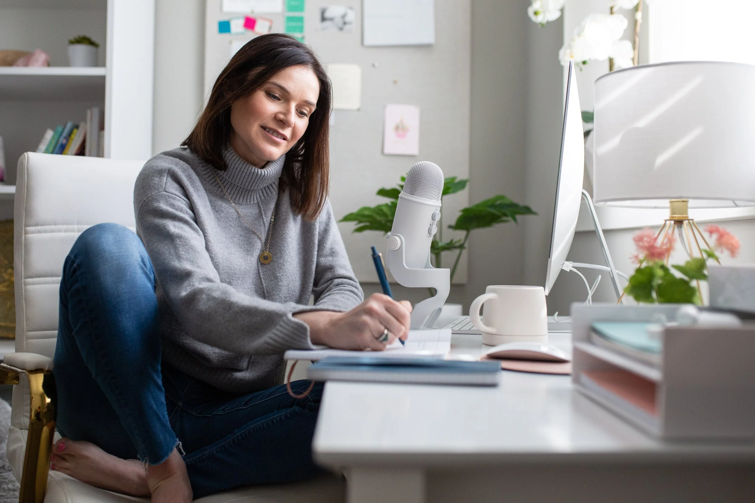 A woman sitting on a white chair at a white desk, writing in a notebook with a blue pen. She is smiling and looking down. On her desk are a white mug, a computer, a microphone, and some documents. Behind her is a bookshelf, a computer monitor, a small plant, a lamp, and some papers pinned on a wall.