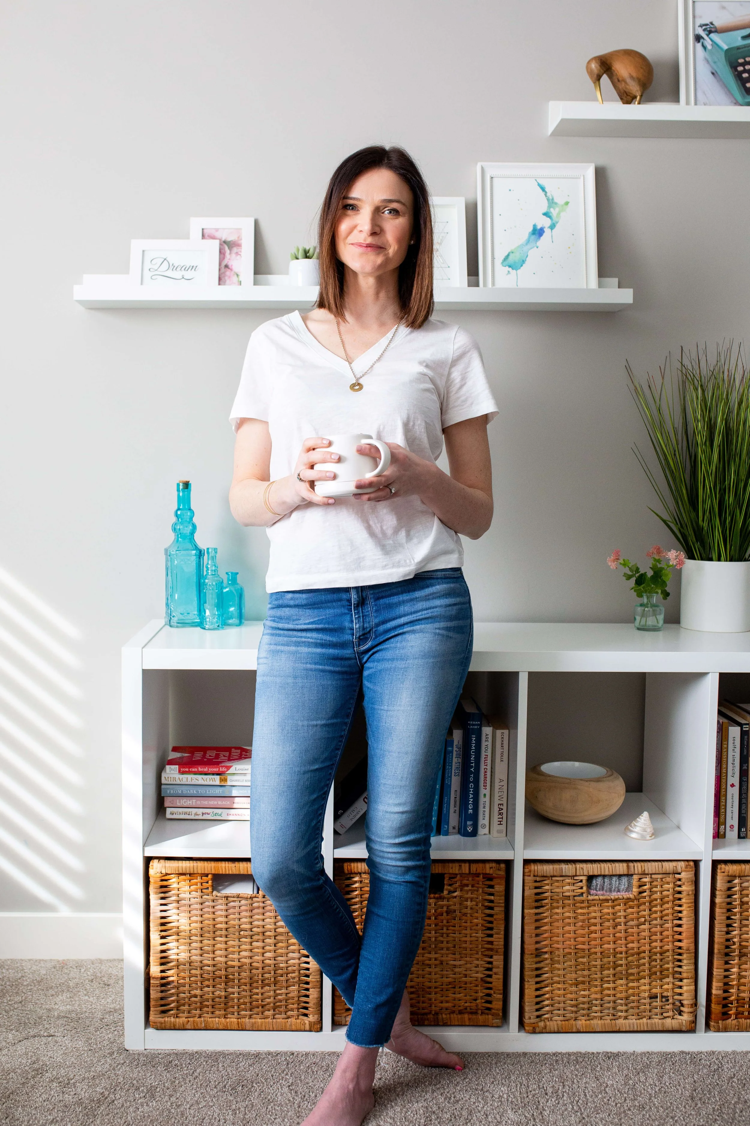 A woman standing barefoot in a room holding a white mug, wearing a white t-shirt and blue jeans, with white shelves, framed artwork, plants, and decorative bottles behind her.