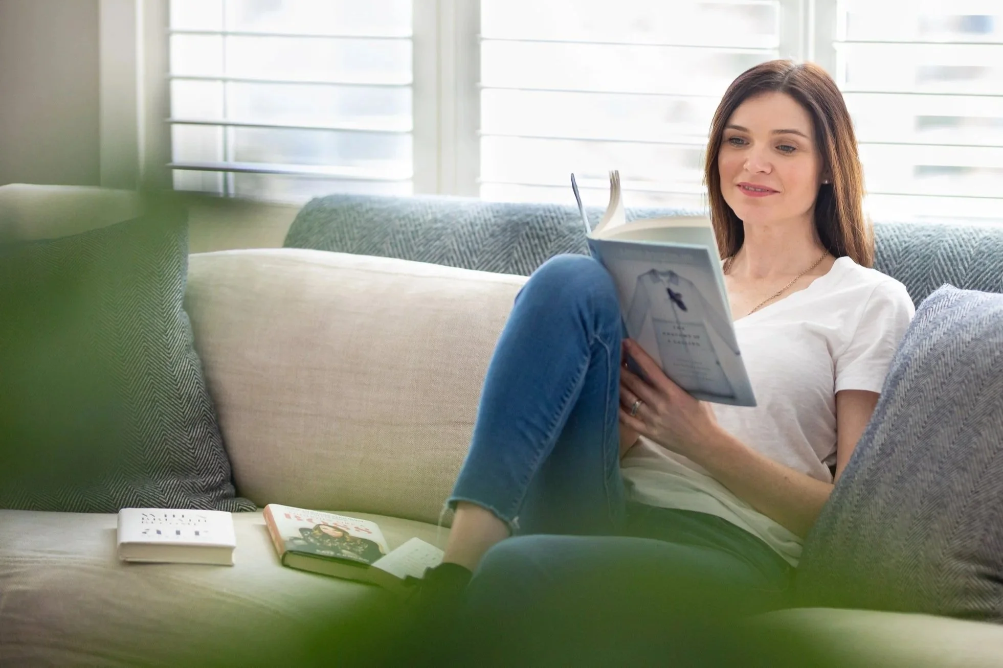 A woman with brown hair in a white shirt and blue jeans sitting on a beige couch, reading a magazine. There are books and a notebook on the couch beside her.
