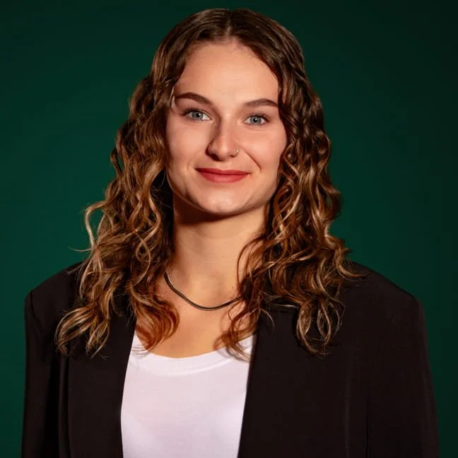 Junge Frau mit lockigem Haar und dunkelblauer Jacke vor grünem Hintergrund.