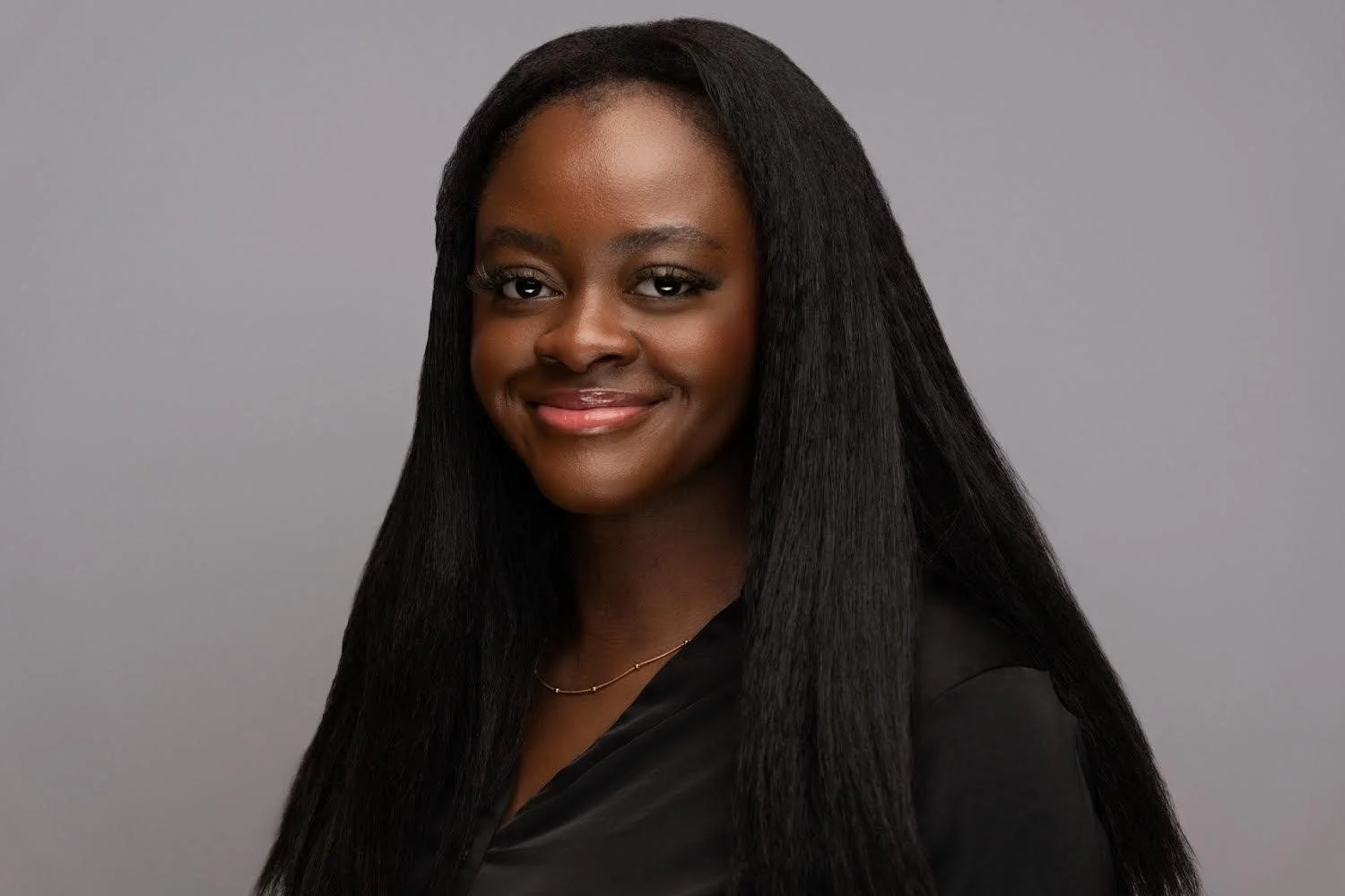 Porträt einer lächelnden schwarzen Frau mit glatten, langen schwarzen Haaren vor grauem Hintergrund.