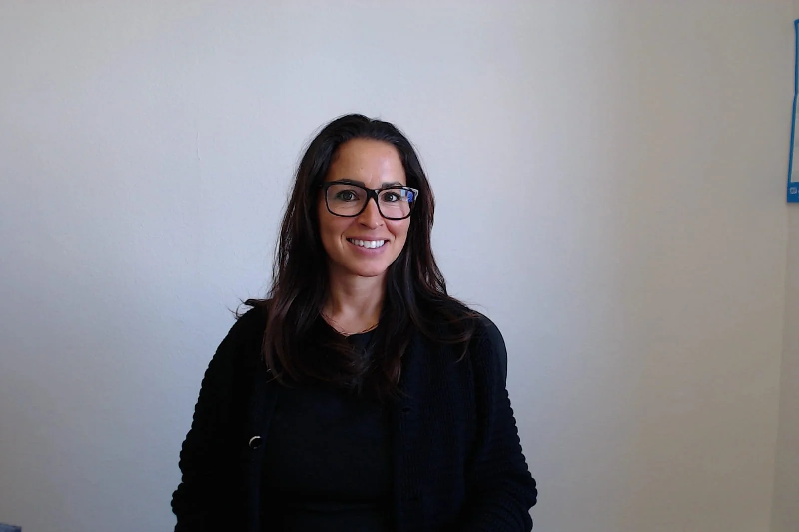 Junge Frau mit Brille und schwarzen Haaren lächelnd vor weißer Wand.