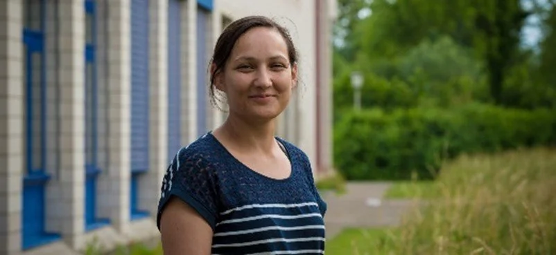 Junge Frau mit braunen Haaren, breiten Lächeln, im blauen gestreiften T-Shirt, im Freien vor einem Gebäude mit blauen Fenstern und grünem Hintergrund, an einem sonnigen Tag.