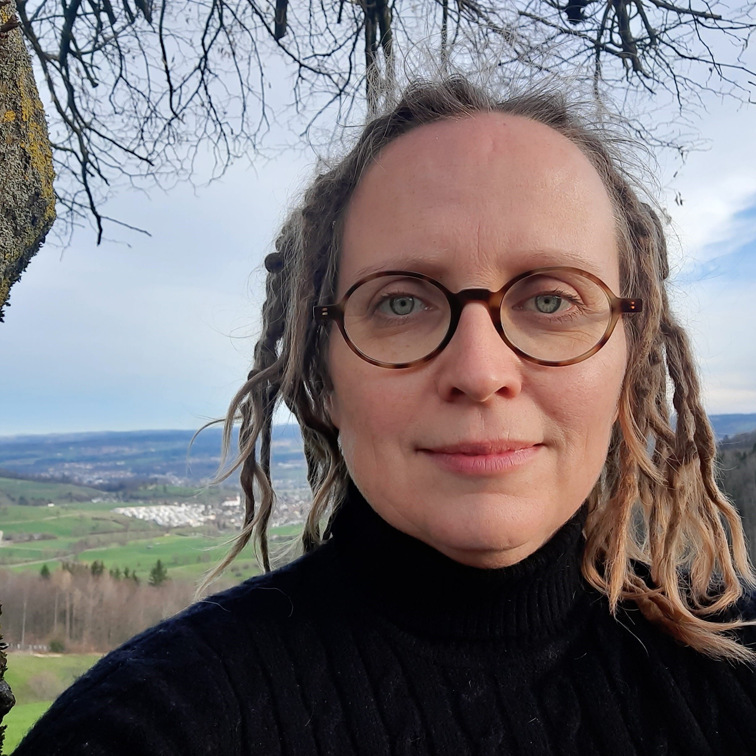 Porträt eines Mannes mit Brille, im Freien, mit Landschaft im Hintergrund, Bäumen und Himmel, wetterbedingt bewölkt.