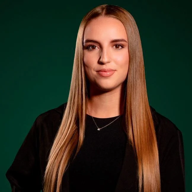 Junge Frau mit glattem, langem, blondem Haar, trägt schwarzes Oberteil und eine kleine Halskette vor grünem Hintergrund.