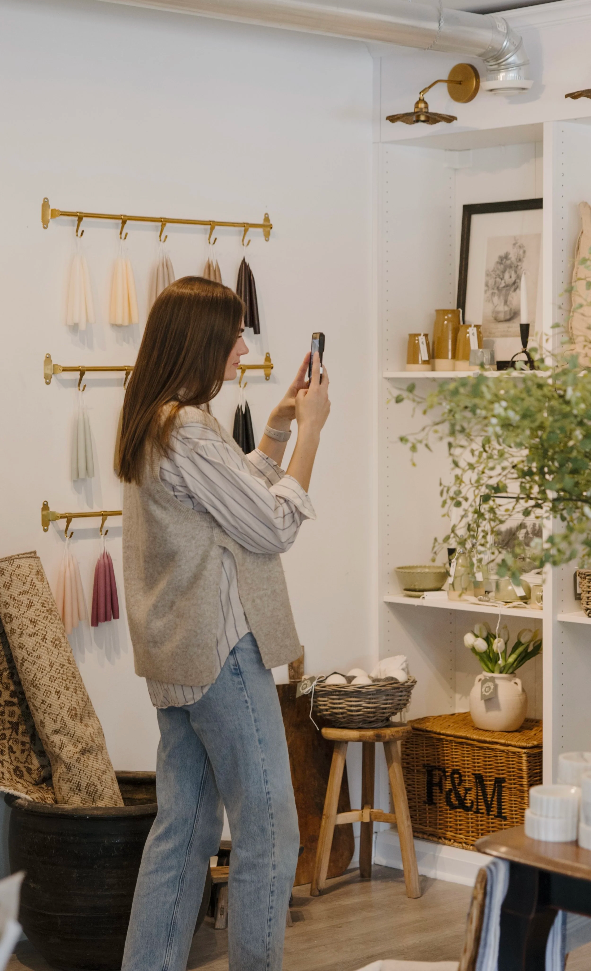 A woman with brown hair, wearing a beige vest over a striped shirt and blue jeans, stands in a decor shop taking a photo with her smartphone. The shop has shelves with home decor items like vases and framed art, and there are fabric swatches hanging on the wall behind her. The display includes woven baskets, plants, and furniture.