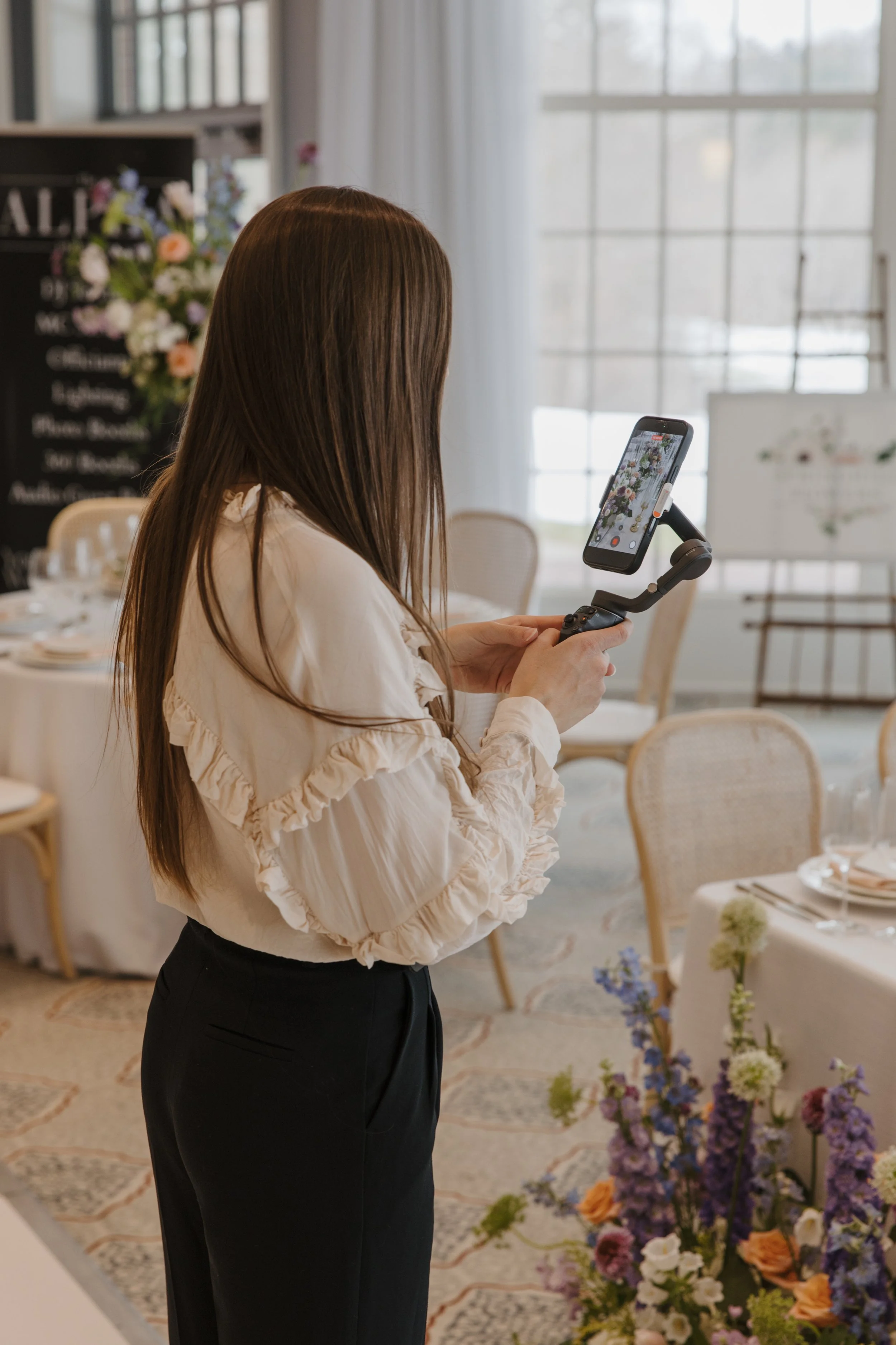 A woman with long brown hair taking a video on an iPhone of a flower arrangement on a wedding or event reception table, in a bright room with large windows and vertical blinds.