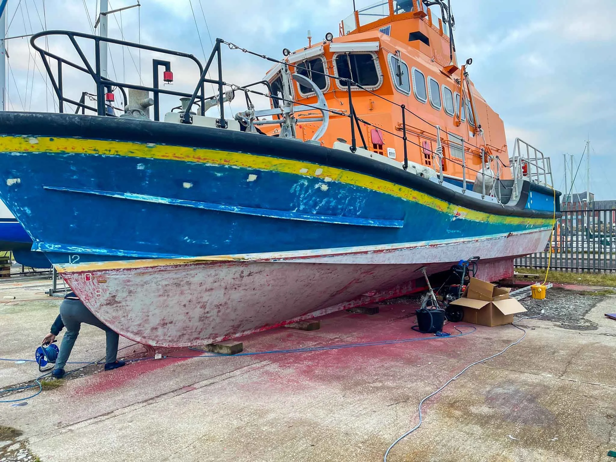 A large boat is being repaired on land with orange superstructure and blue hull, propped up on blocks, surrounded by tools, cords, and a person working underneath.