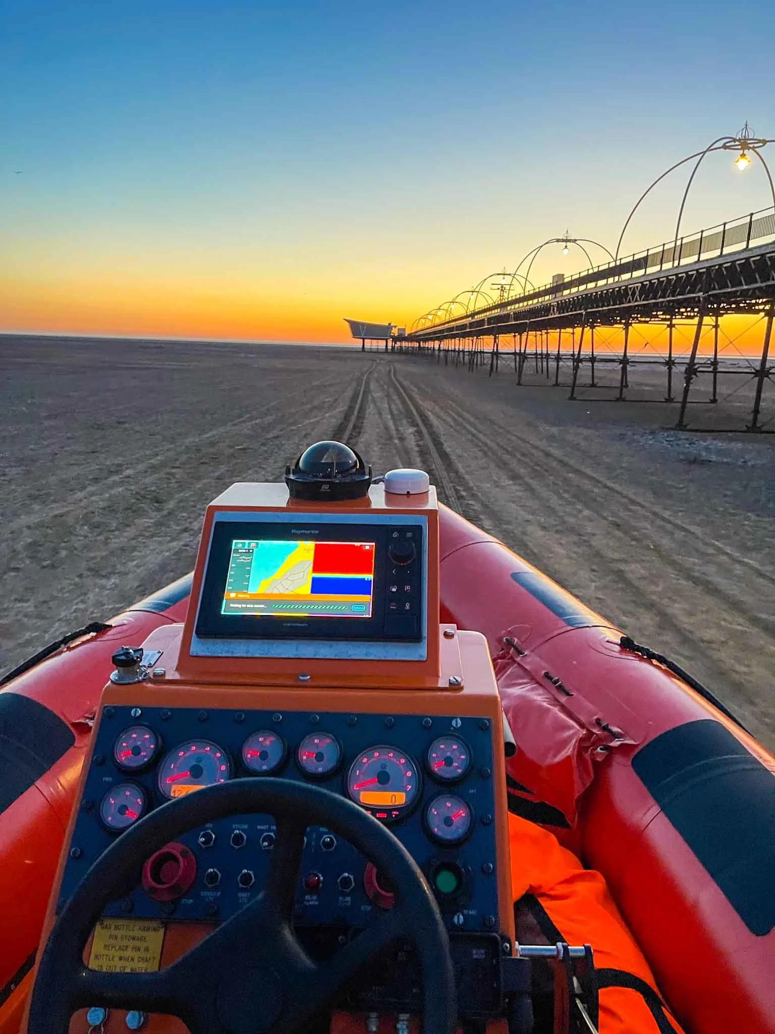 View from inside a rescue boat on the beach at sunset, with a navigation screen visible and a pier extending into the ocean.