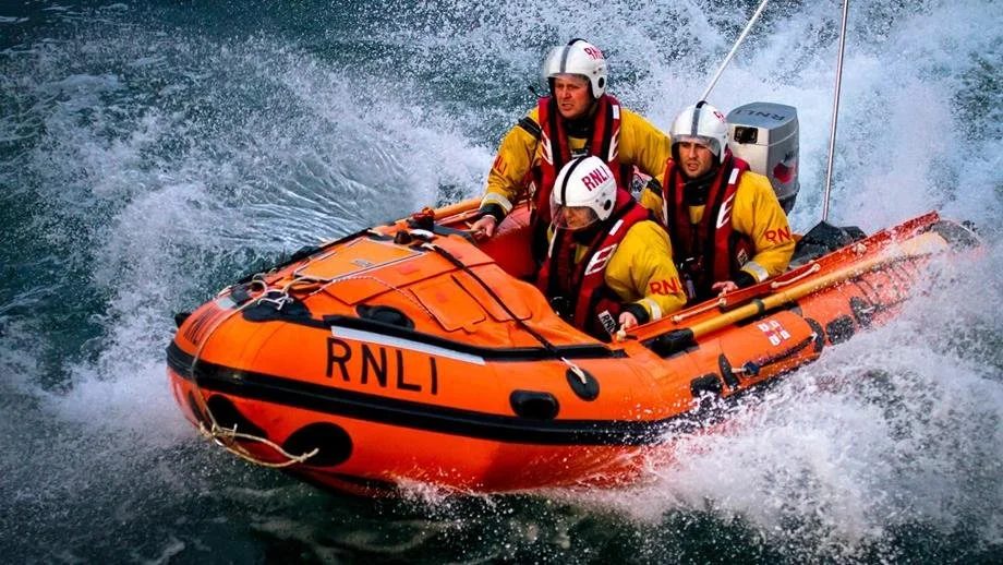 Four rescue team members wearing helmets and yellow jackets riding an orange boat on rough water.