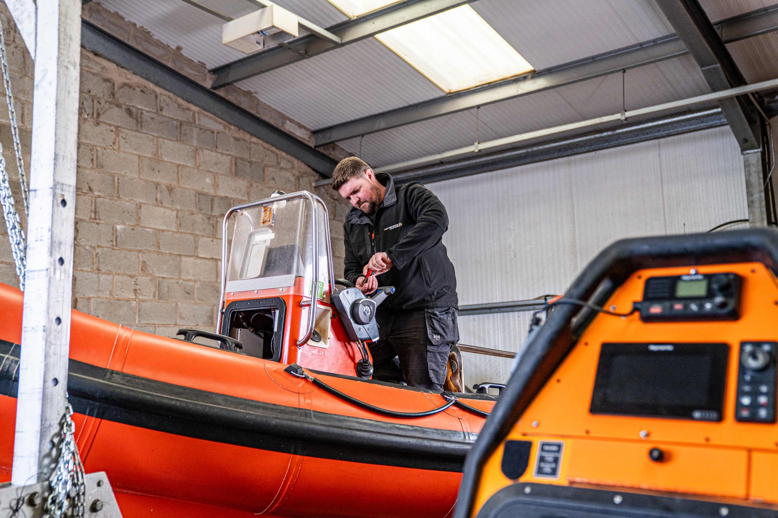 A man working on an orange rescue boat inside a warehouse, wearing a black jacket, with equipment and control panels around him.