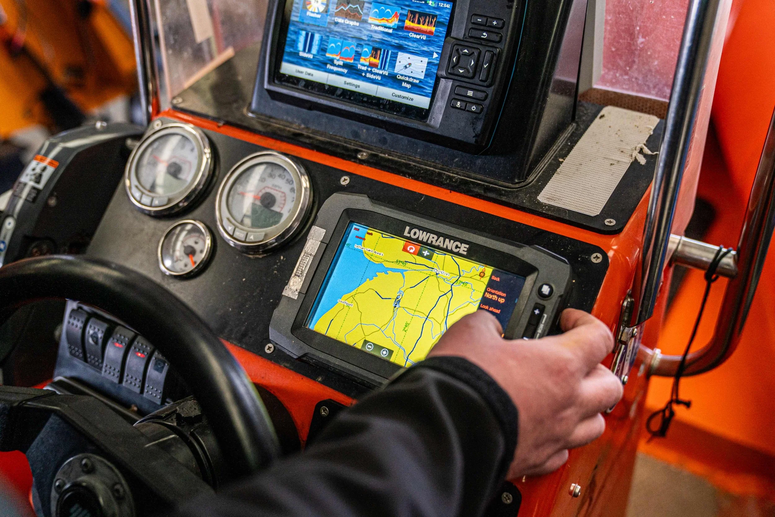 A smiling woman with shoulder-length brown hair, wearing round glasses, small triangle earrings, and a black top, standing indoors with a light-colored wall and green plant in the background.