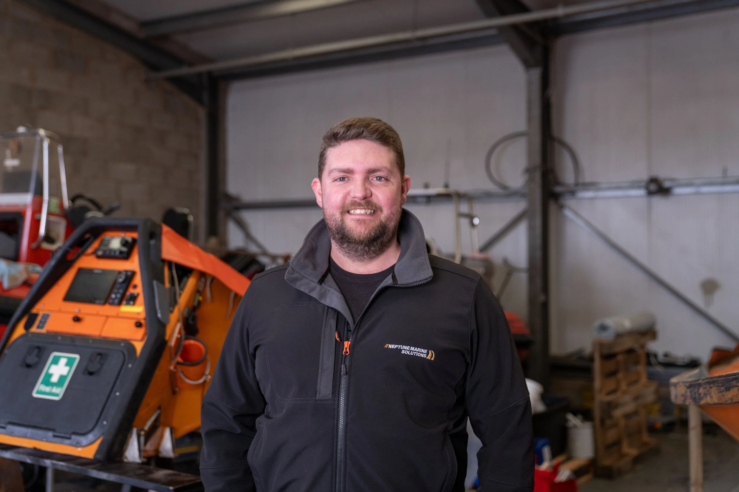 A man with a beard and short brown hair wearing a black jacket standing in a warehouse with equipment and tools in the background.