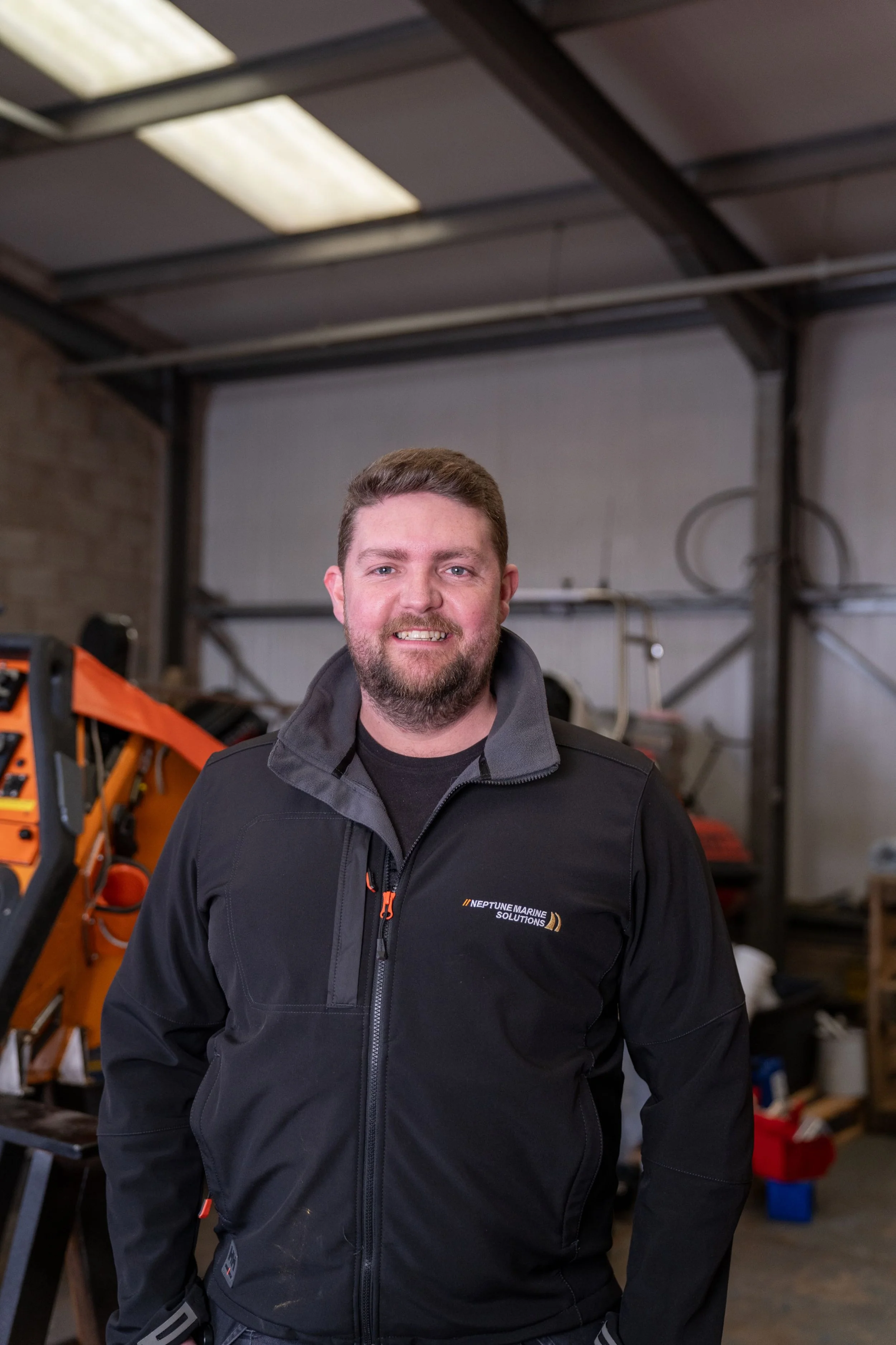 A smiling man with a beard wearing a black jacket, standing inside a workshop with equipment and tools in the background.