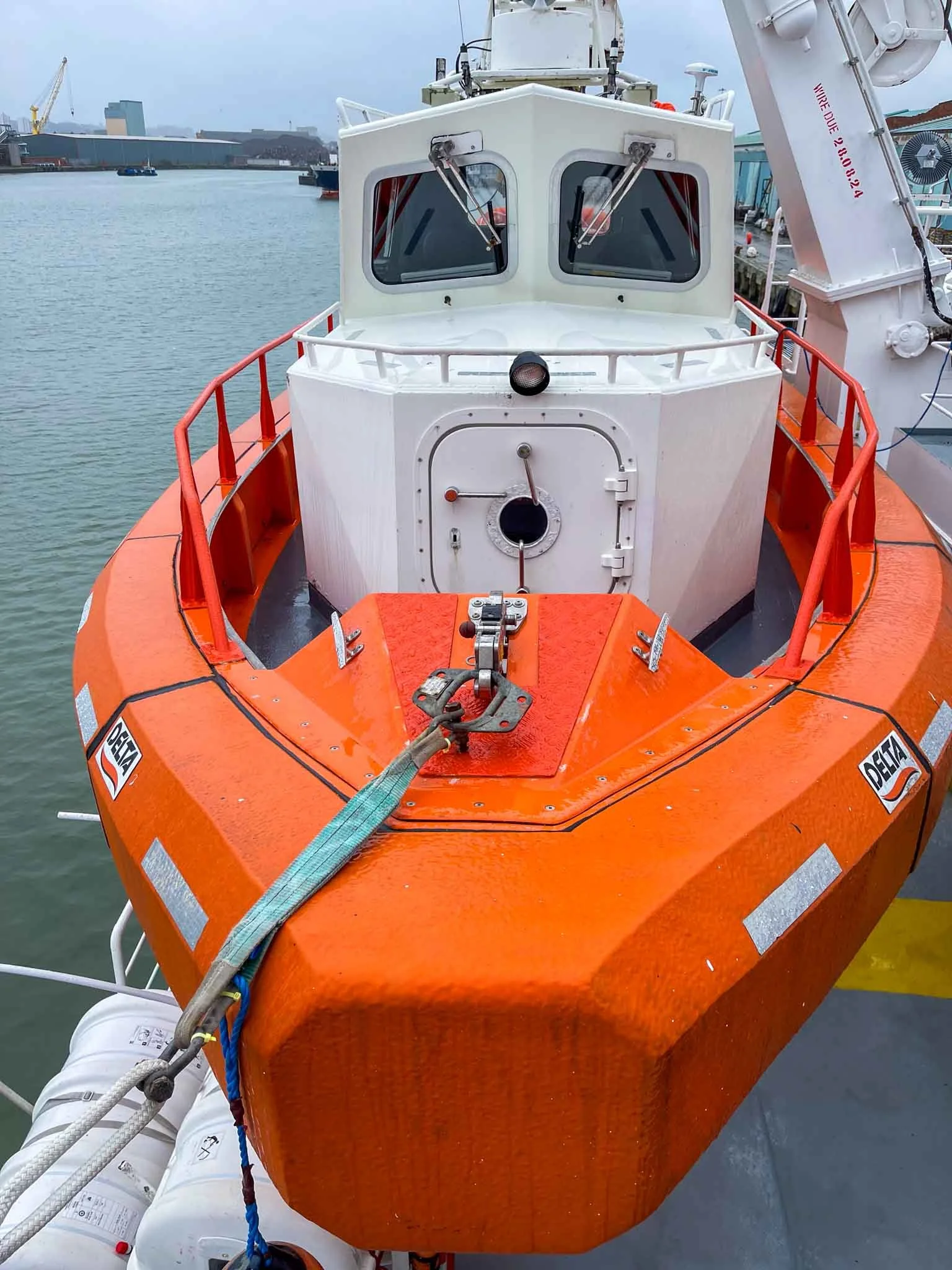 Front view of a boat with an orange hull docked at a marina, showing the bow with a round hatch, windowed control cabin, and safety equipment attached.