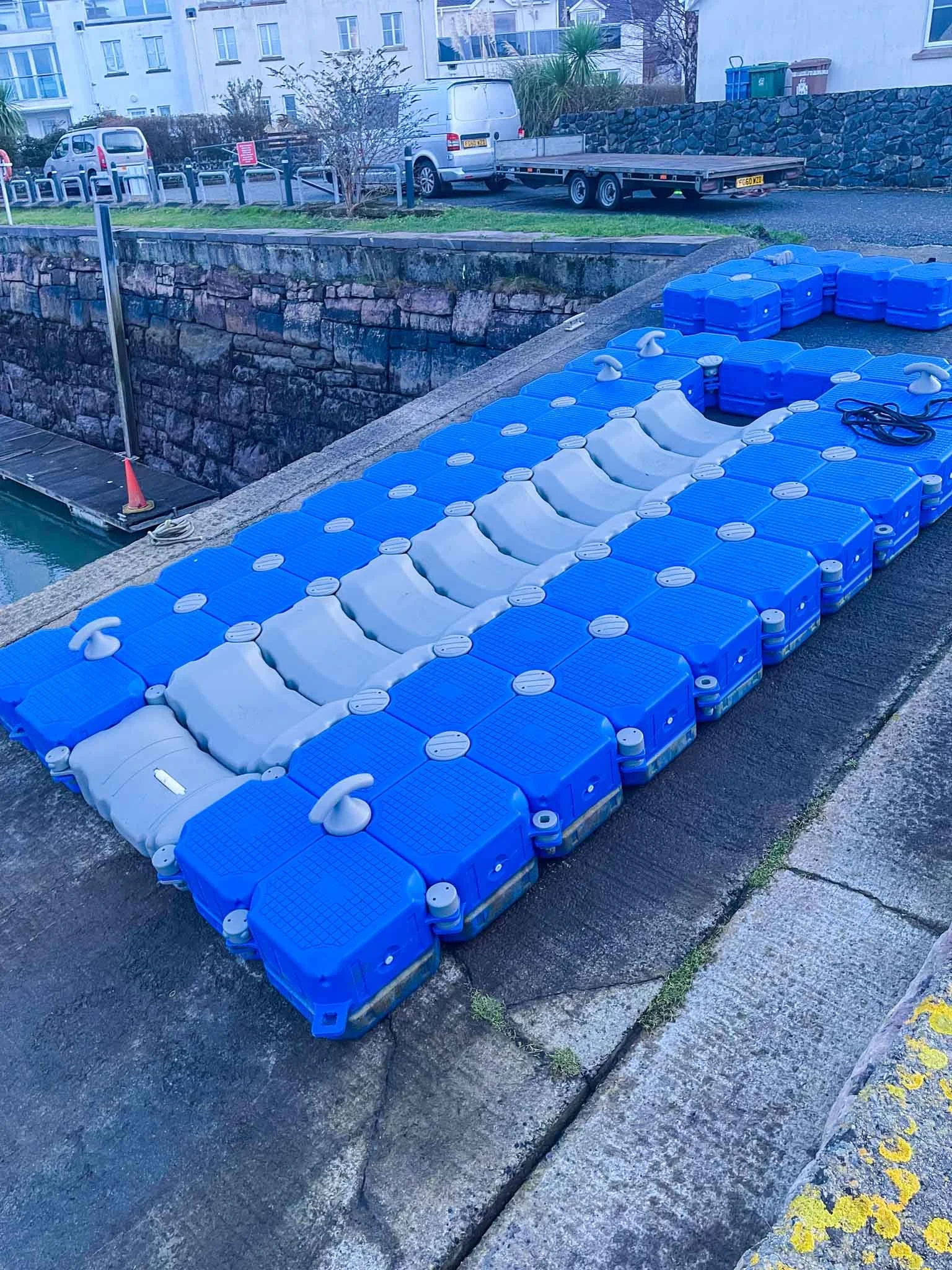 Several blue and gray floating plastic docks and storage containers stacked by a canal or harbor, with a trailer and parked vehicles in the background.
