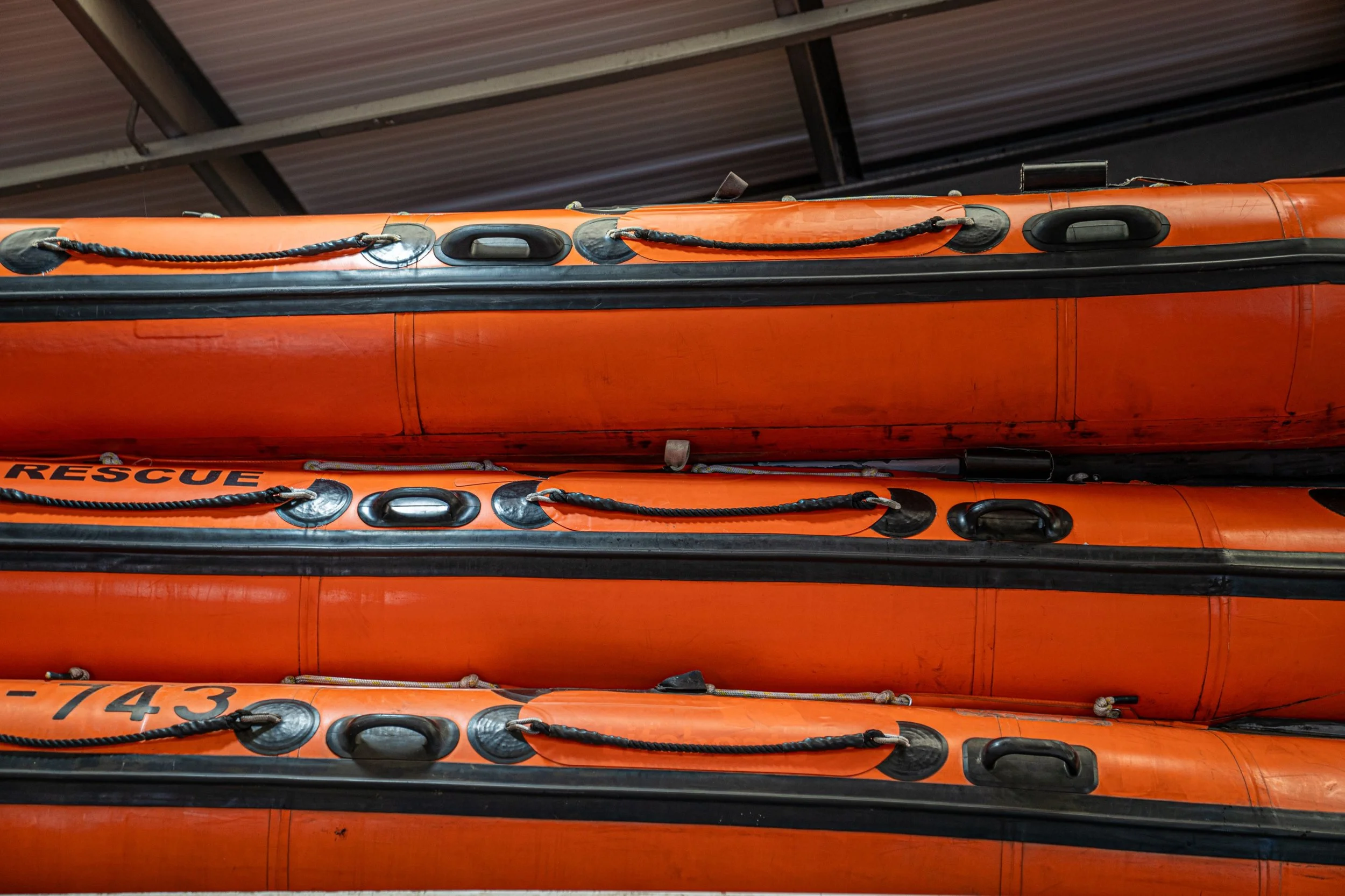A stack of orange rescue boats with black handles and ropes, stored upside down under a metal roof.