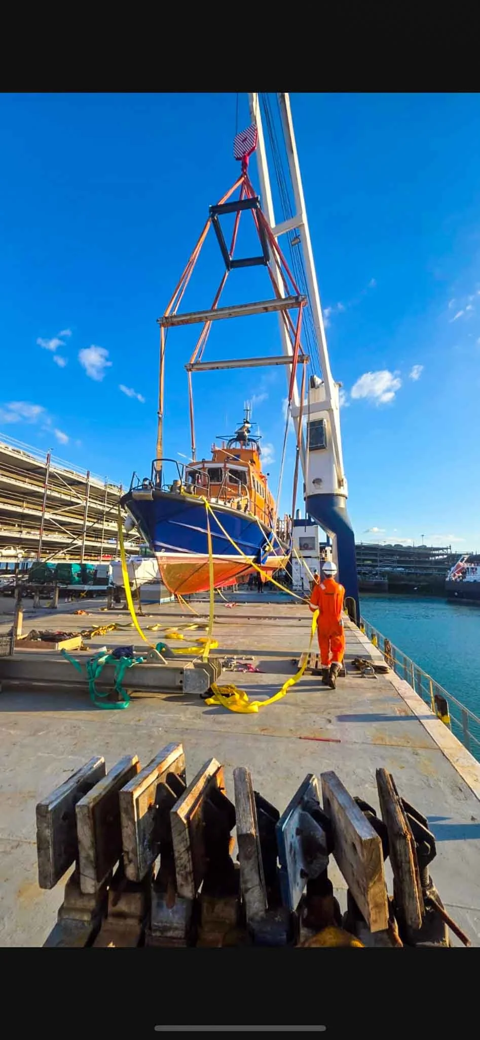 A rescue worker in orange uniform walking on a dock, guiding a boat lifted by a crane, with ropes and equipment around, under a clear blue sky.
